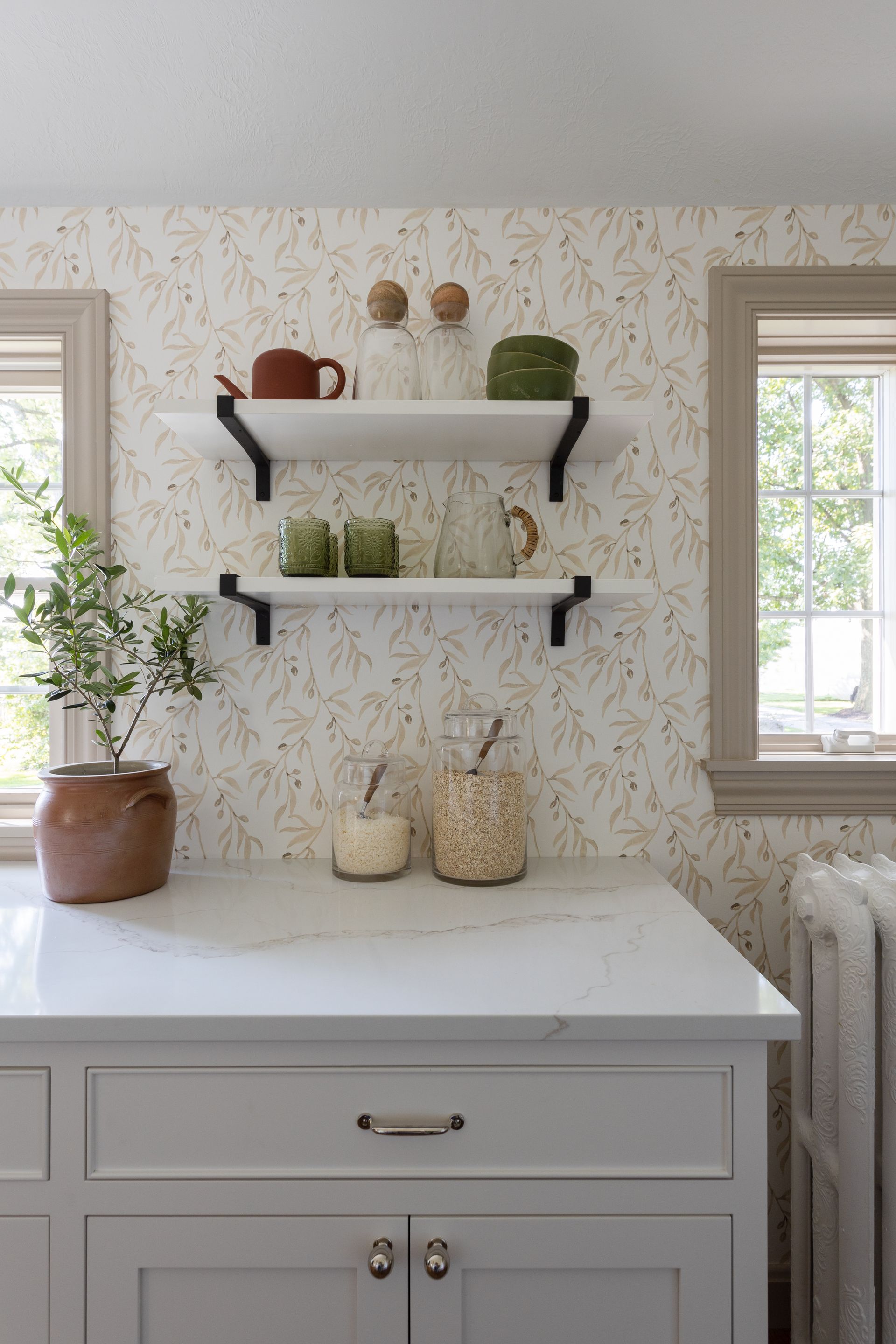 Kitchen with shelves, wallpaper, and countertop. Displayed items include jars, plants, and dishes.