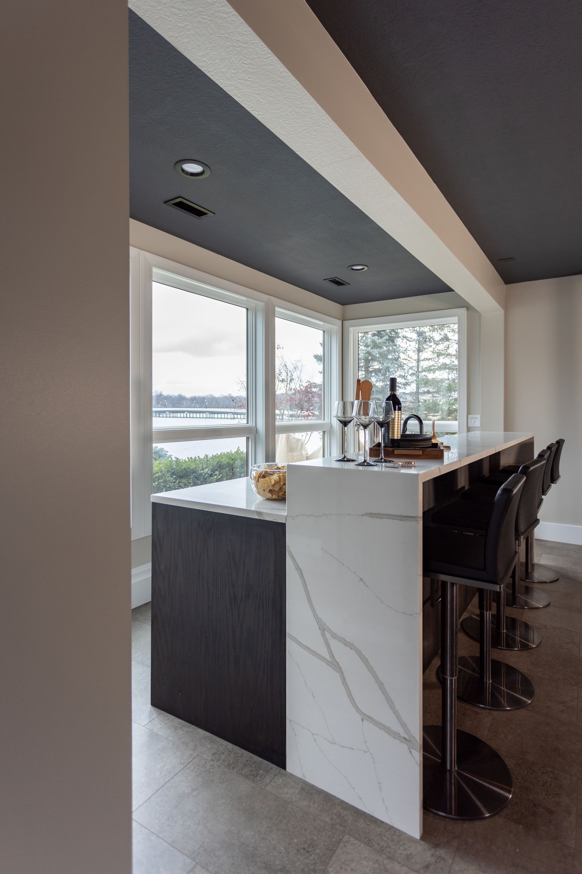 Bar with white marble countertop, dark wood base, and black bar stools, near a window with a water view.