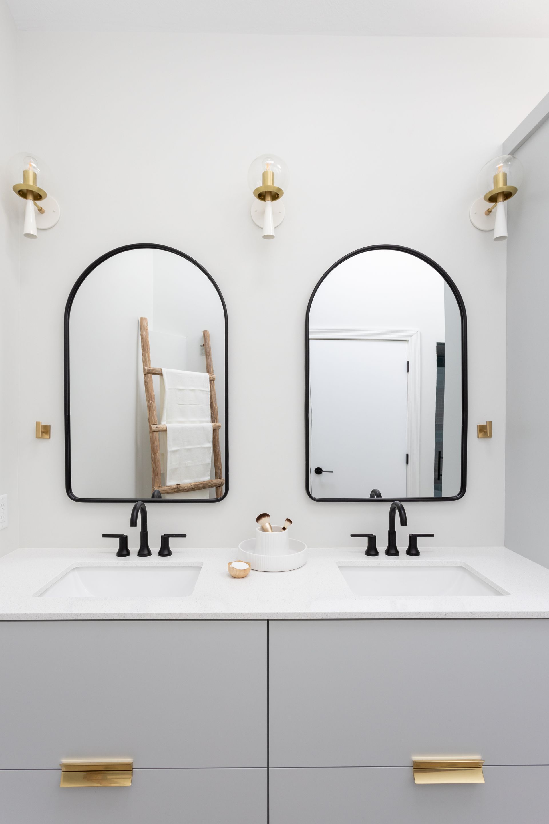 Bathroom vanity with gray cabinets, black arched mirrors, gold hardware, and sconces.