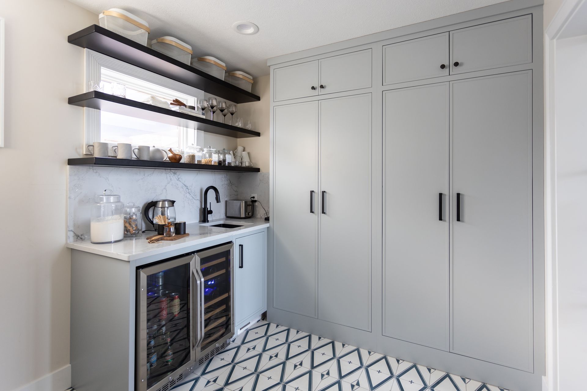 Gray pantry with built-in cabinetry, mini fridge, and sink. Black shelves above, patterned floor.