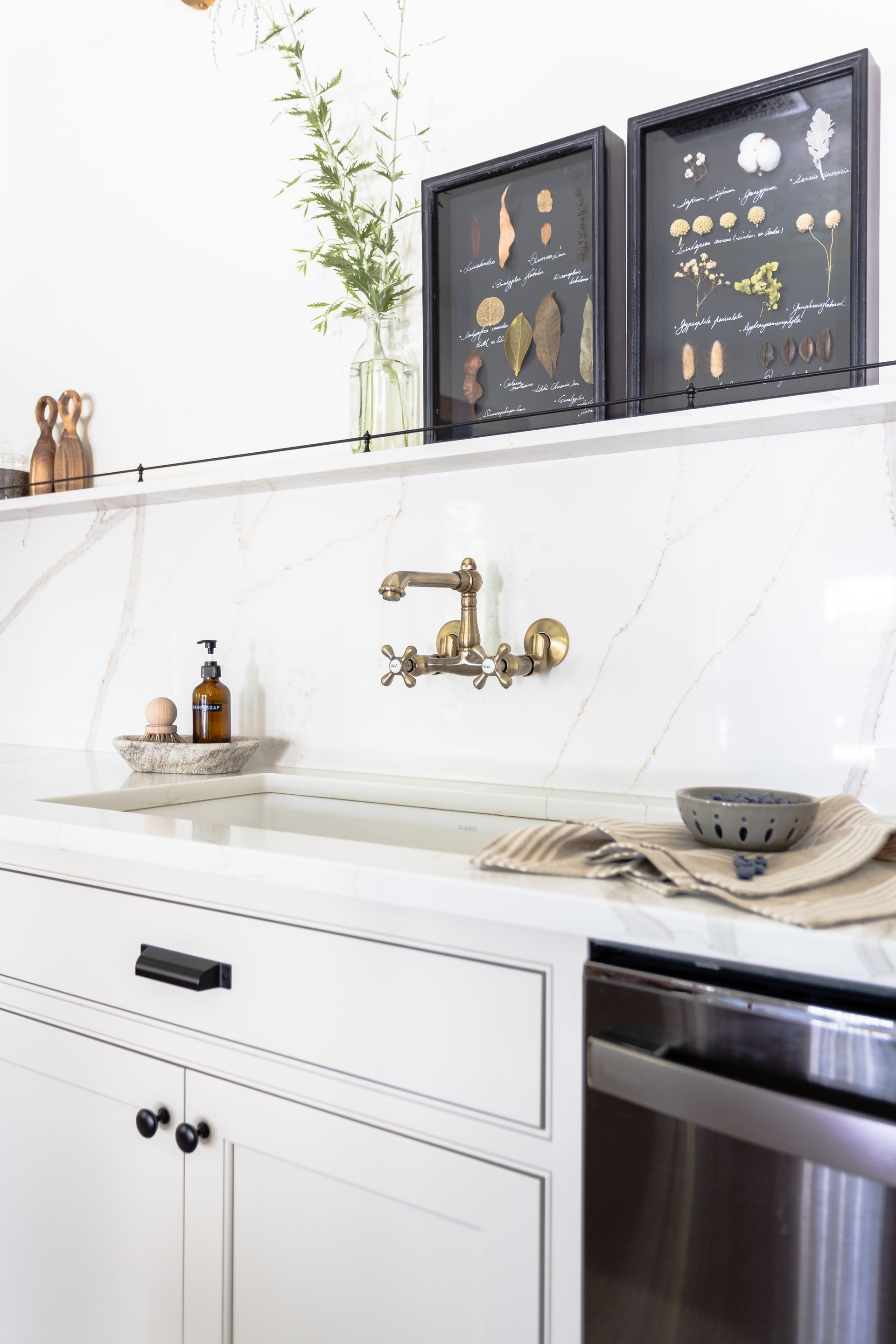 White kitchen with marble backsplash, brass faucet, and framed botanical art.