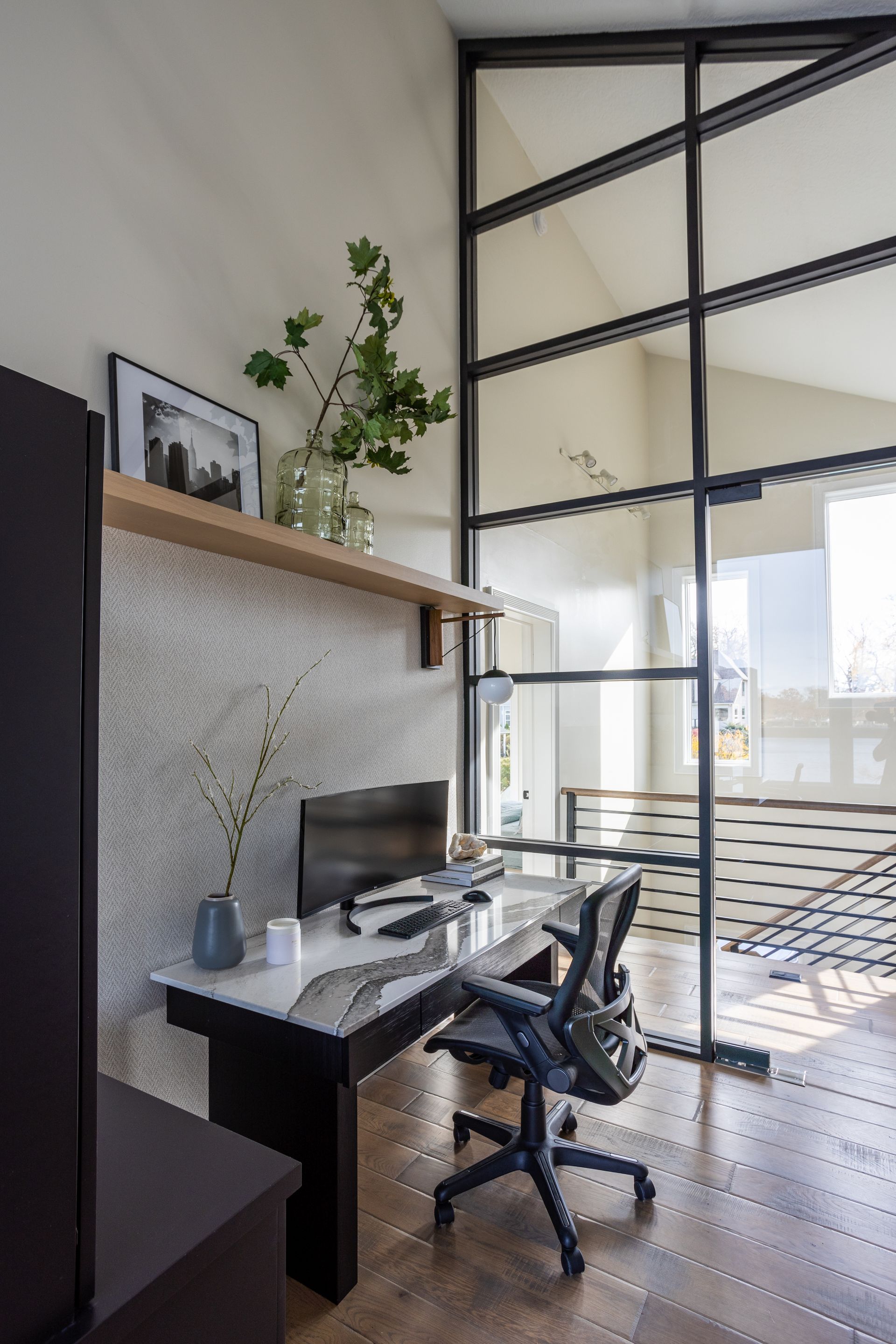 Home office with desk, computer, and chair. Tall window with black frame. Beige and white walls, wood floor.