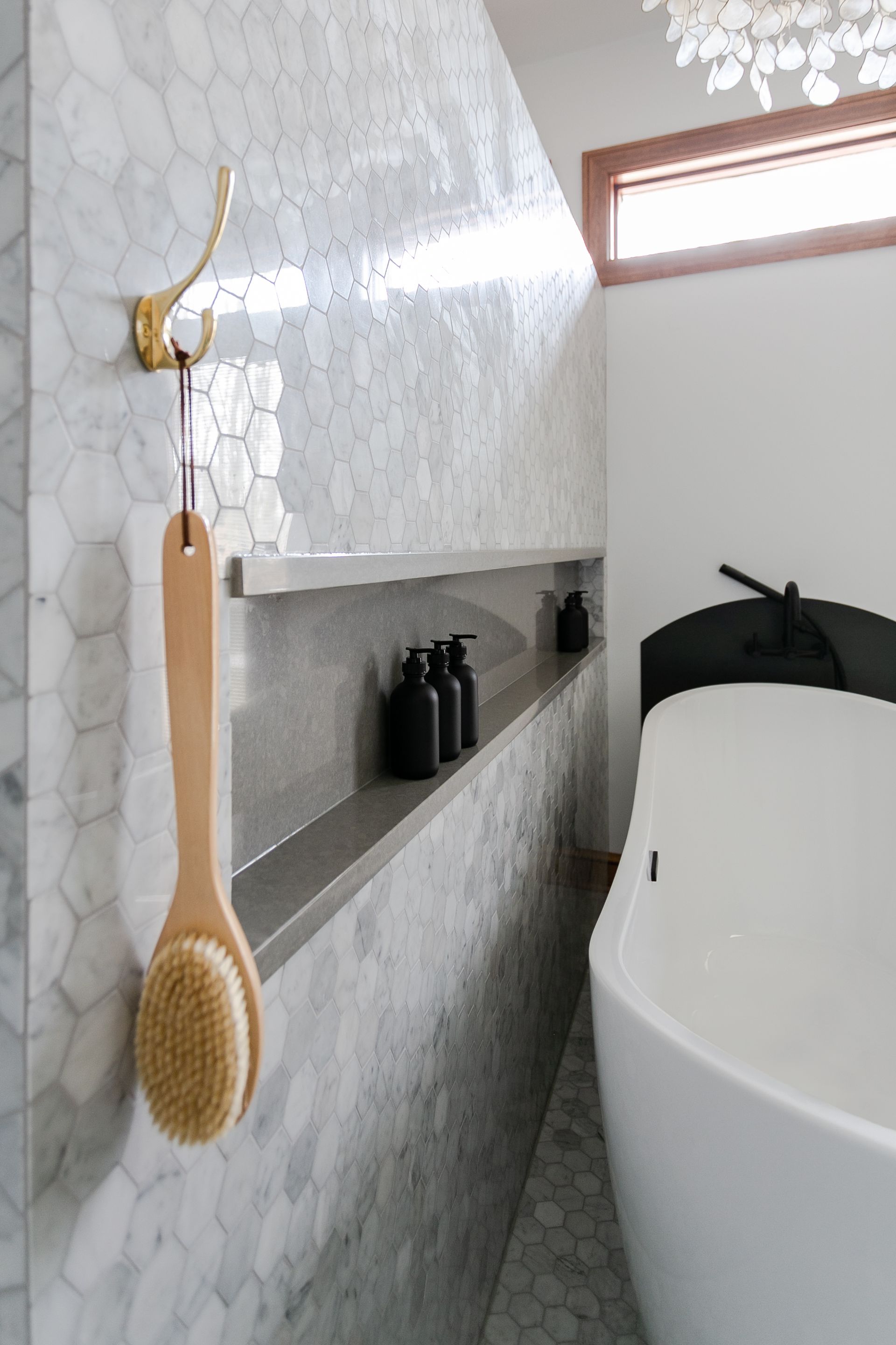 Bathroom with marble tile walls, built-in shelf with bottles, a soaking tub, and a hanging brush.