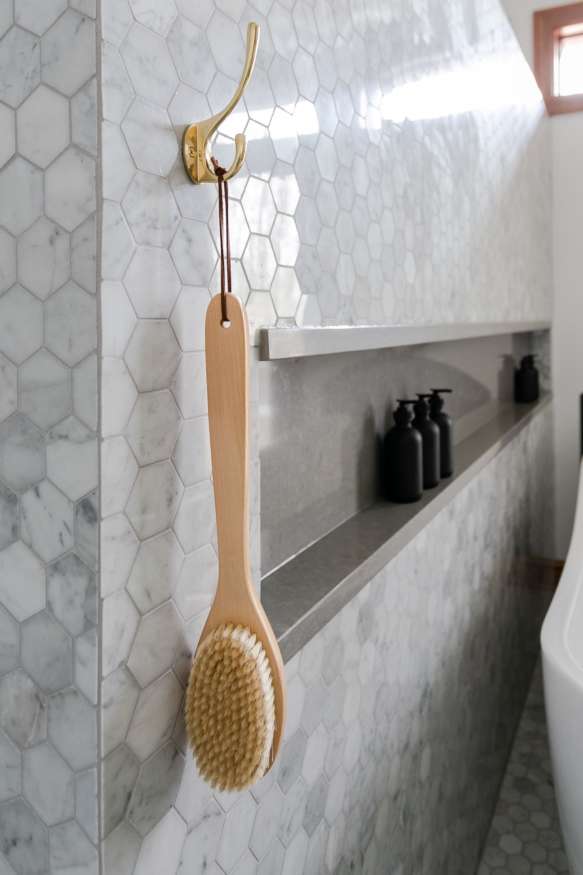 Wooden body brush hanging on a gold hook in a tiled bathroom with a shelf holding black bottles.