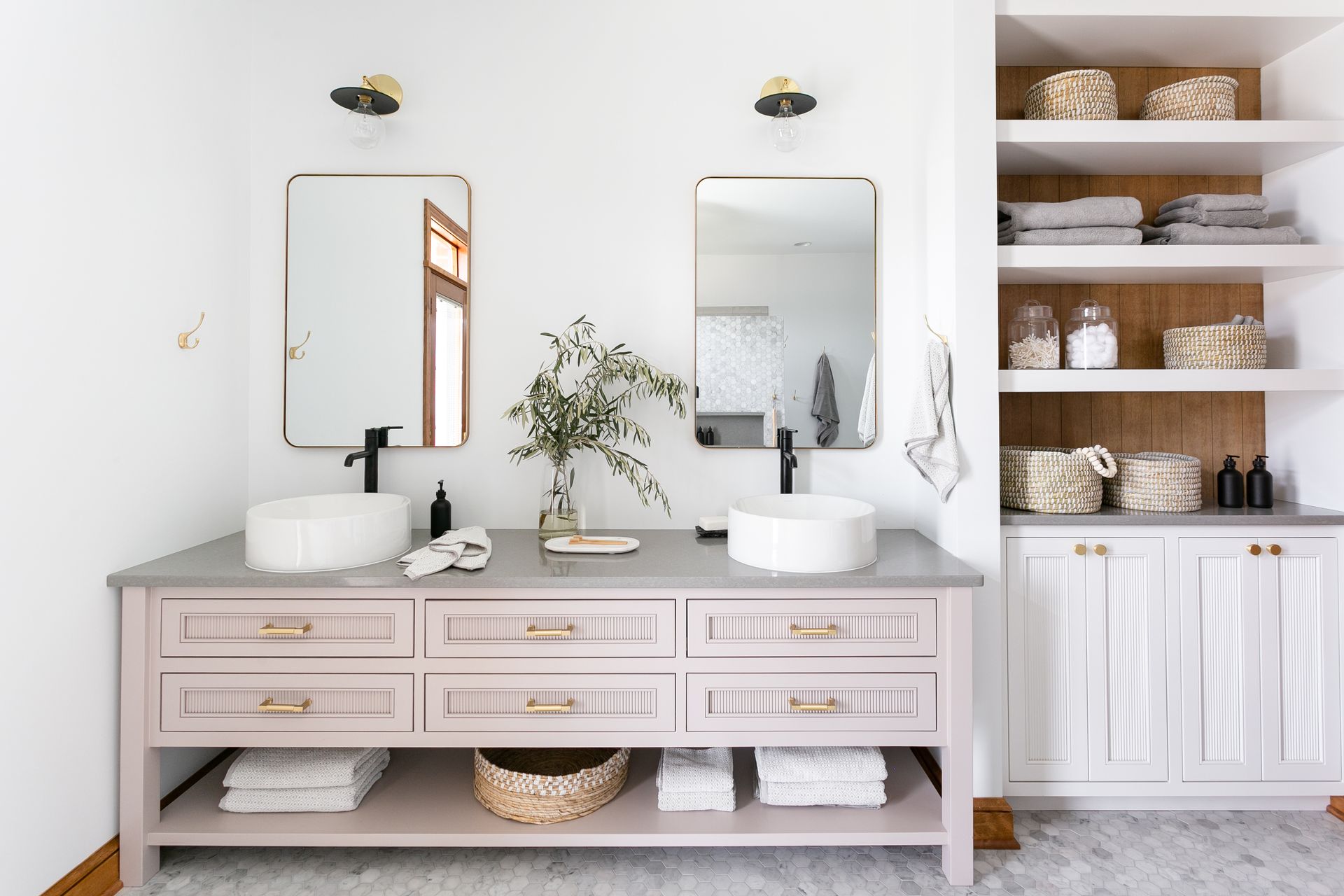 A bathroom with double sinks, mirrors, and open shelving. Pink vanity with gray countertop, white walls.