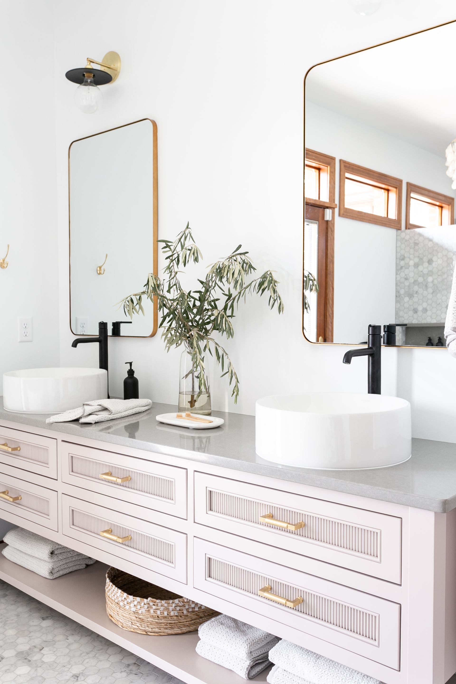 Modern bathroom with double vanity, gold-trimmed mirrors, black faucets, light pink cabinets, and a vase of greenery.