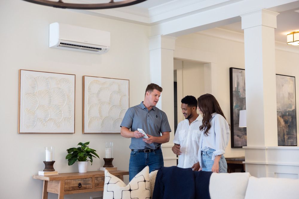Three people talking in a bright living room with white walls and a wall-mounted air conditioner.