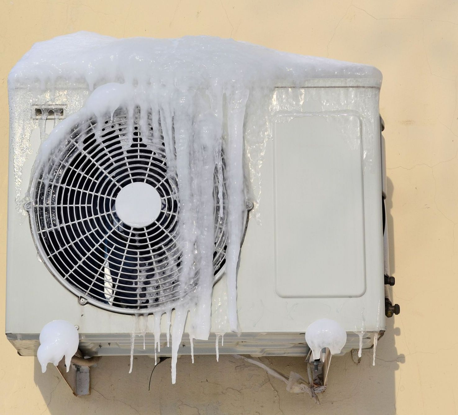Outdoor air conditioner covered in thick ice and icicles on a beige wall