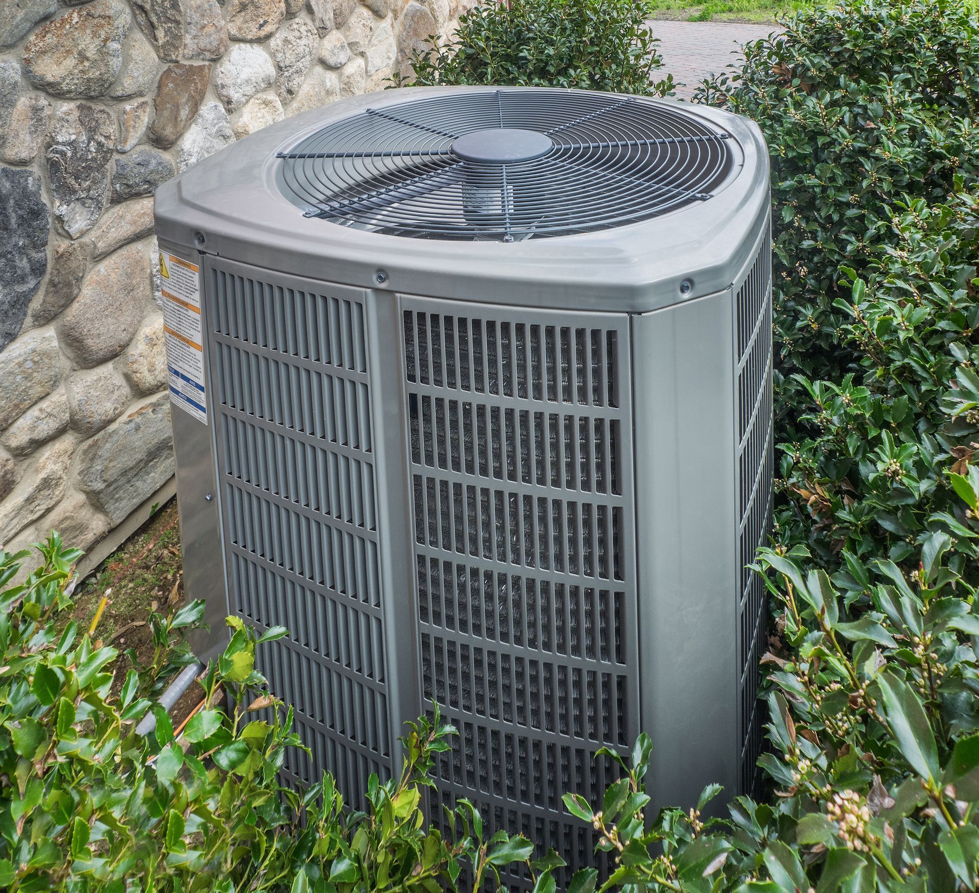 Outdoor air conditioner unit beside a stone wall, partially surrounded by bushes