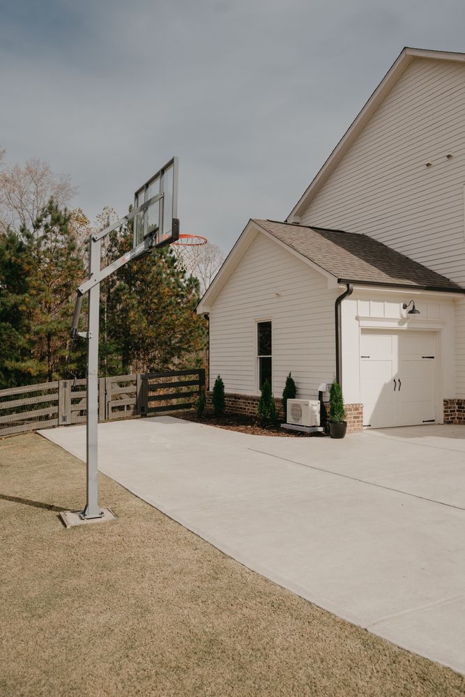 Suburban driveway with a white house, garage, basketball hoop, and fenced yard under cloudy sky