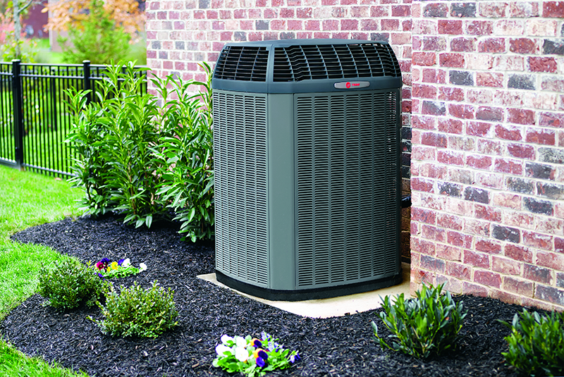 A large air conditioner is sitting on the side of a brick building. Outdoor HVAC unit beside a brick house wall, surrounded by shrubs and mulch in a landscaped yard