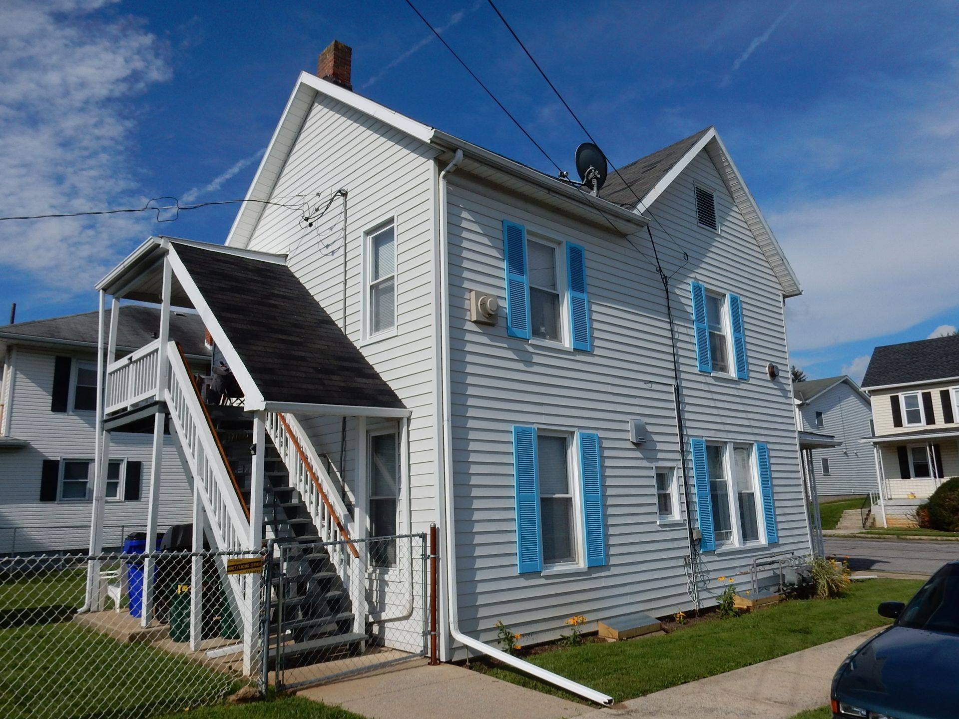 White two-story house with blue shutters and an exterior staircase on a sunny street corner