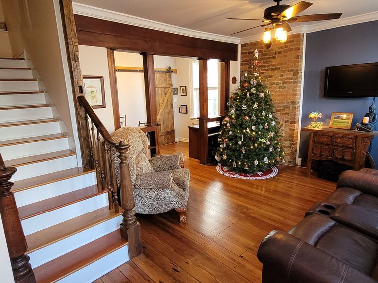 Cozy living room with staircase, armchair, leather sofa, and lit Christmas tree by a stone fireplace.