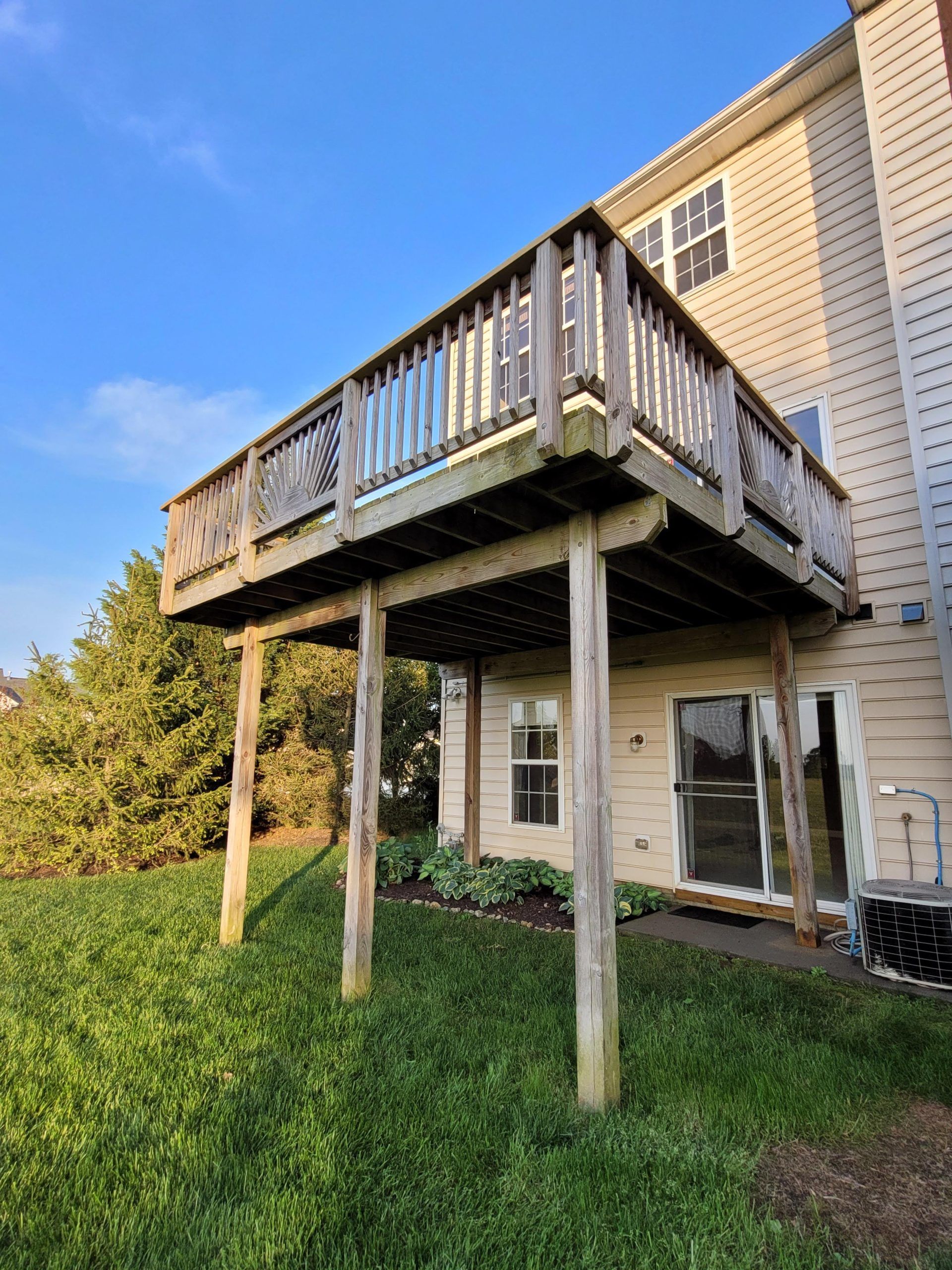 Elevated wooden deck attached to a beige house, with stairs, posts, and a grassy backyard below.