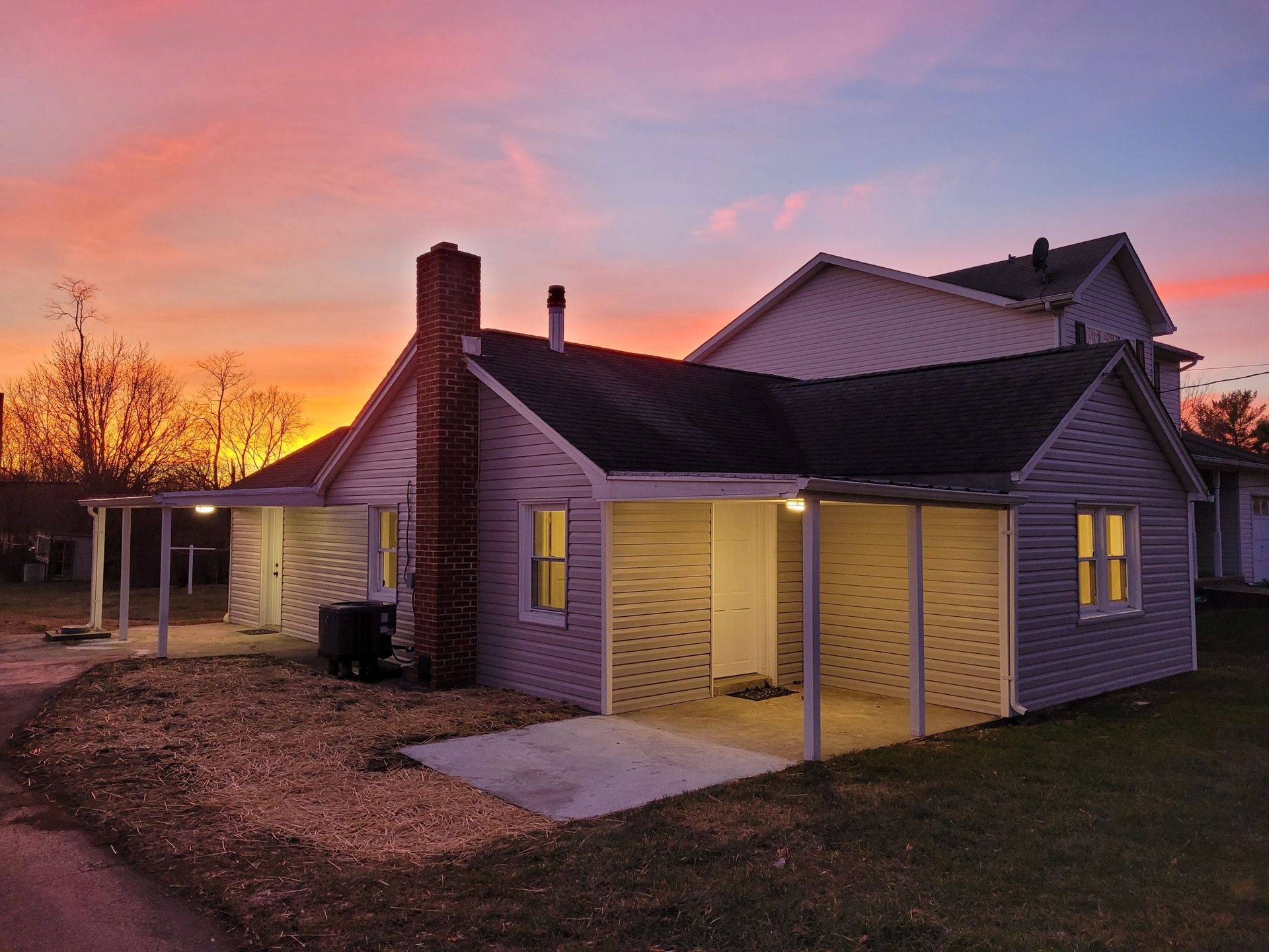 Small house with lit porch at sunset under a pink-orange sky