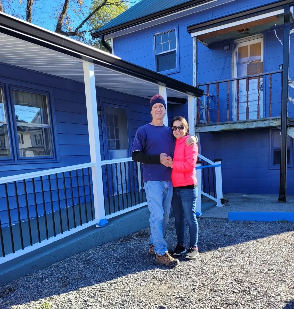 Two people posing outside a blue house with a porch in bright sunlight
