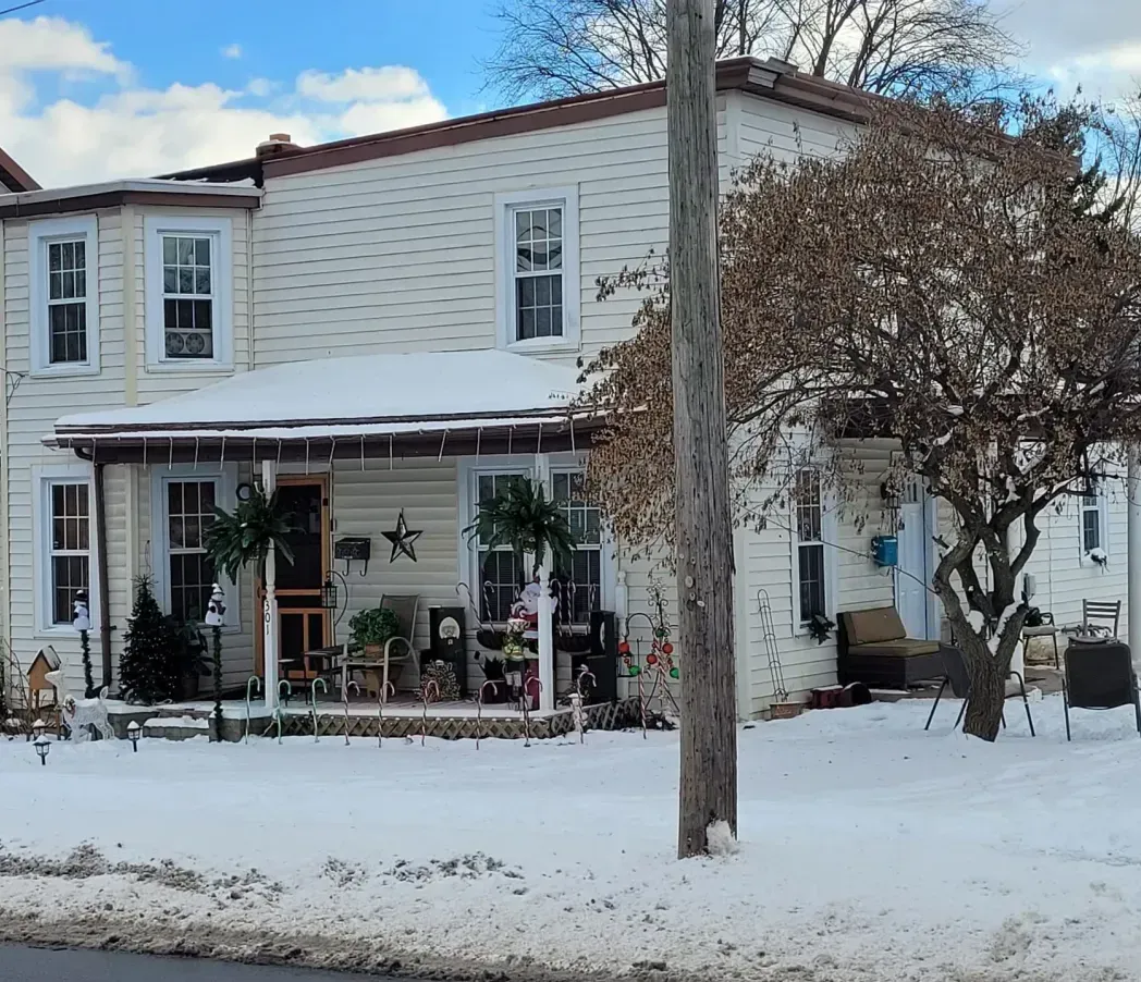 Snow-covered two-story house with a porch, windows, and a large tree in front