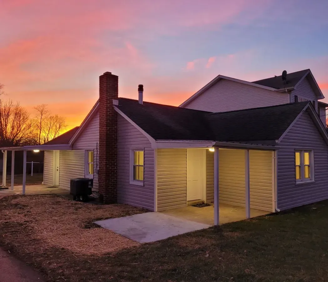 Small purple house with lit porch at sunset under a pink-orange sky.