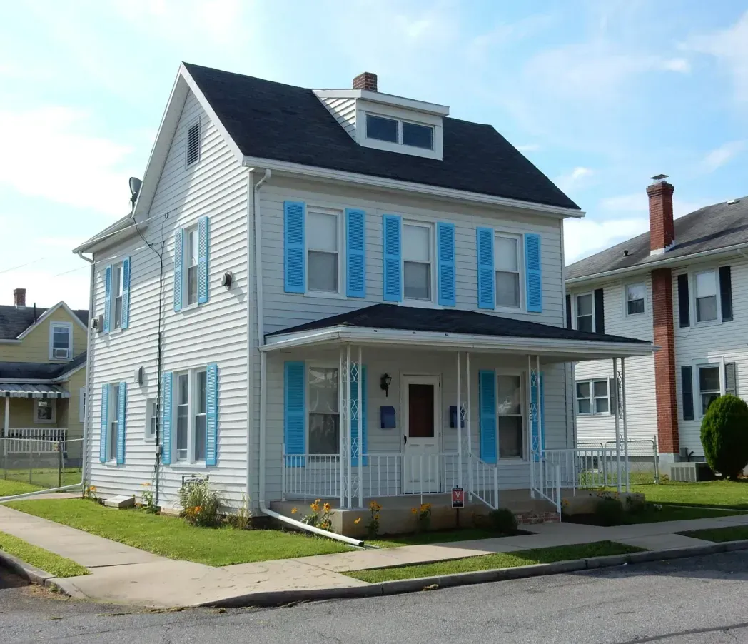 White two-story house with blue shutters and porch, on a sunny residential street