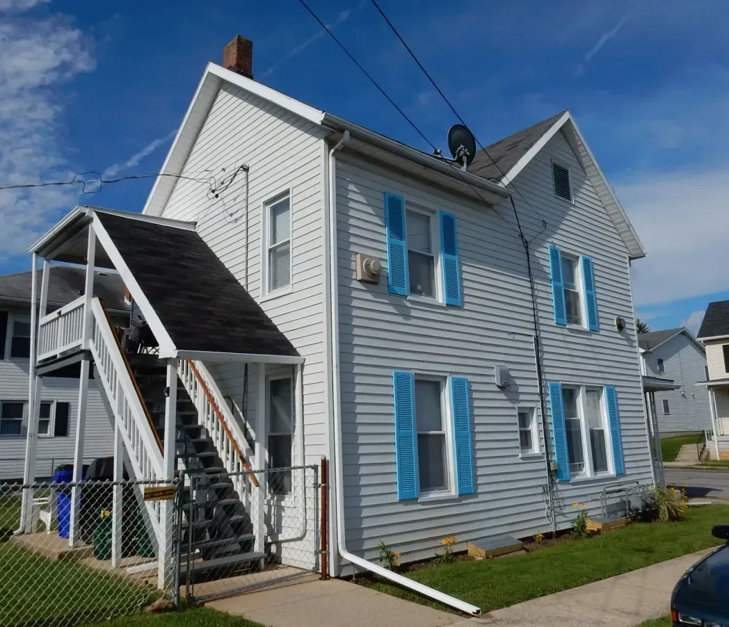 White two-story house with blue shutters and an exterior staircase on a sunny street.