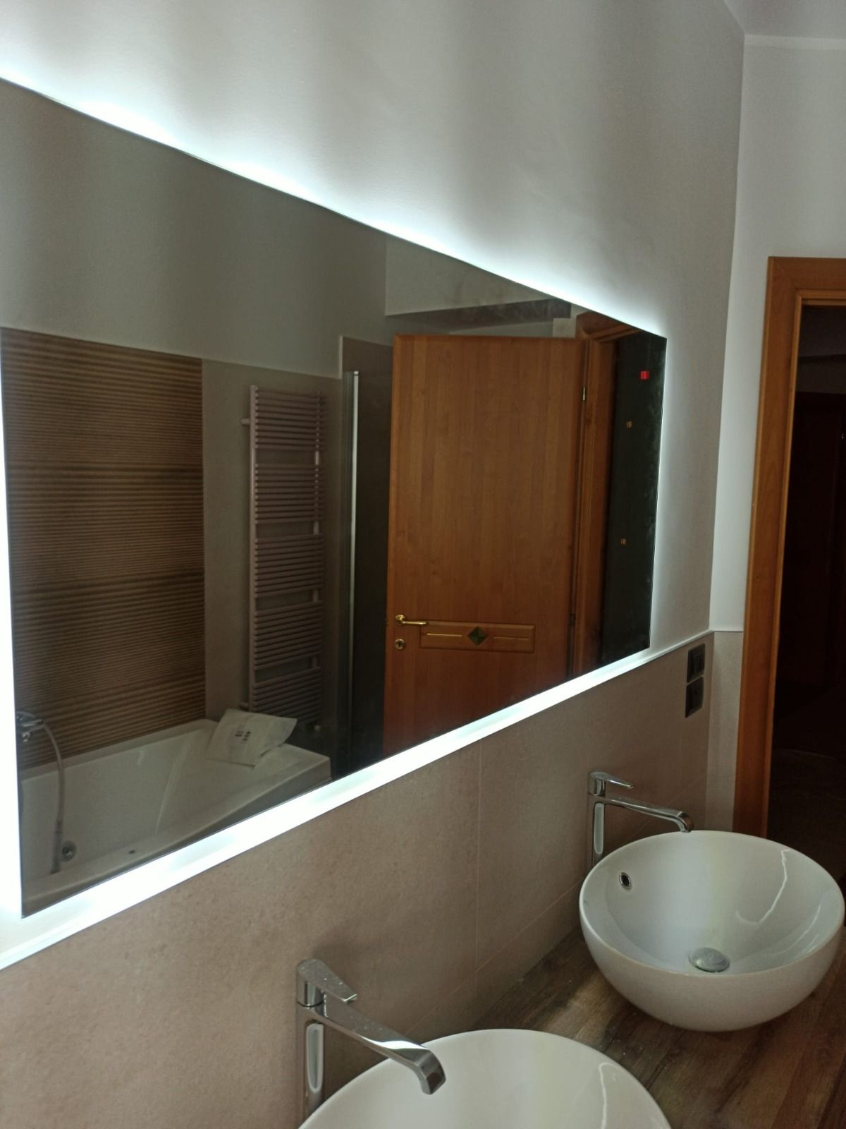 Bagno moderno con grande specchio retroilluminato sopra due lavandini. Include vasca, doccia e porta in legno.