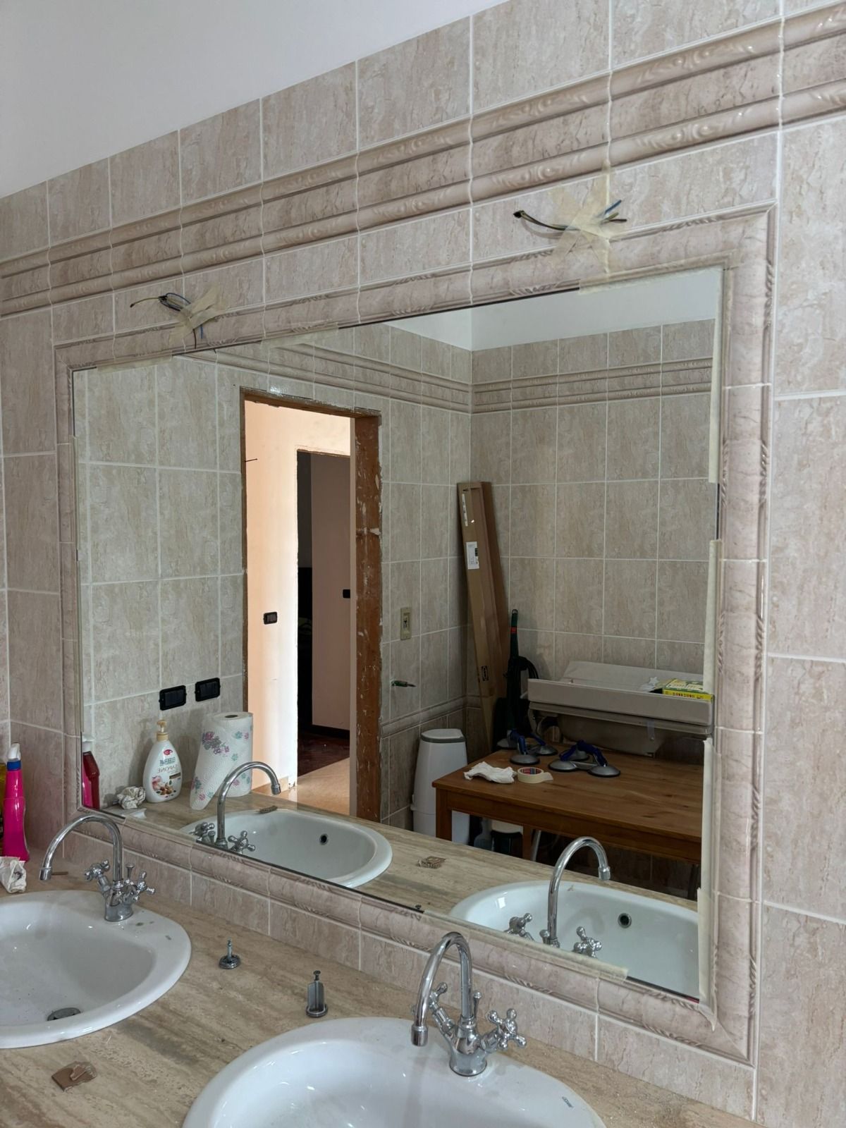 Bagno con grande specchio sopra due lavandini. Pareti piastrellate beige, mobiletto e tavolo in legno visibili nel riflesso dello specchio.