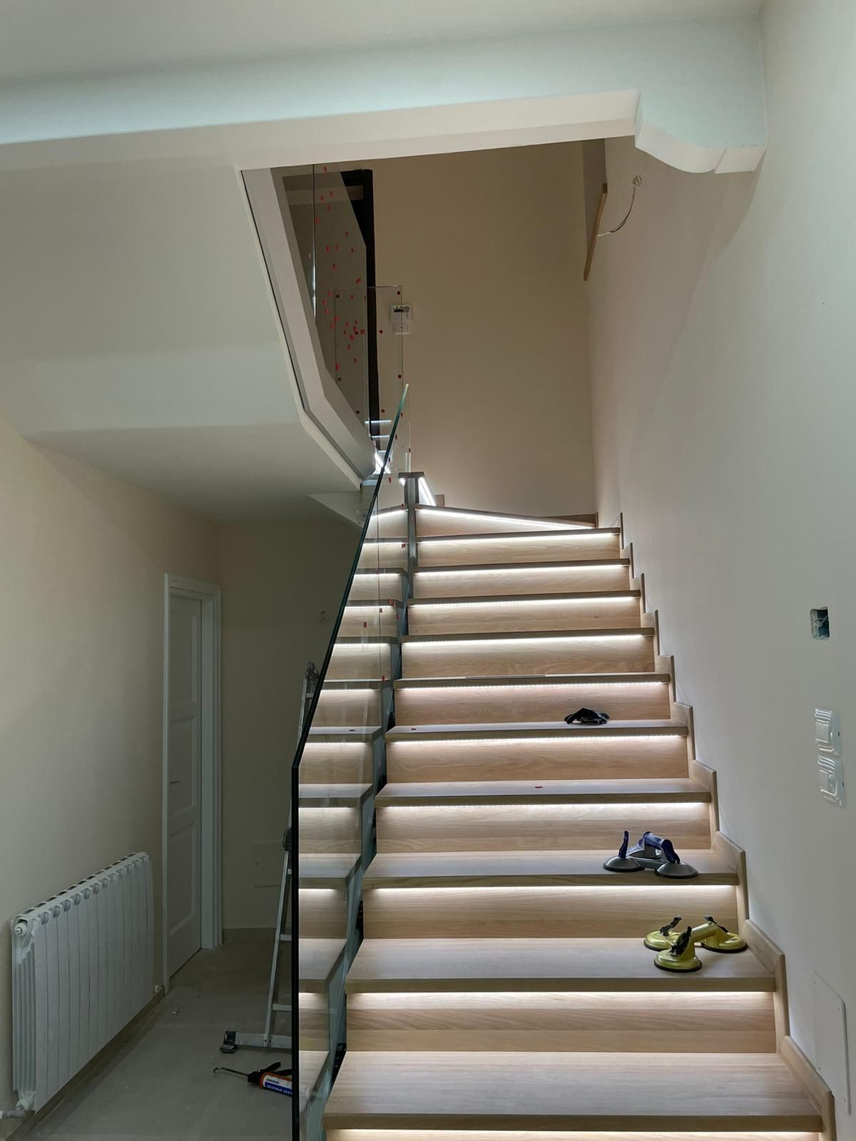 Scala con gradini in legno e ringhiera in vetro. Ogni gradino è illuminato. Pareti beige.