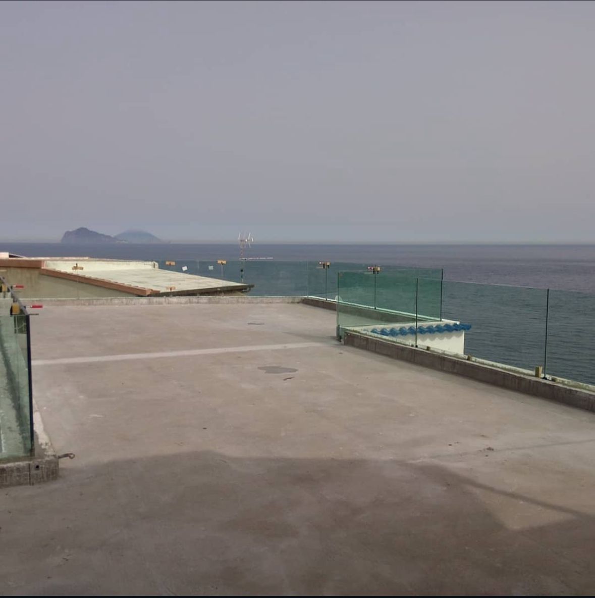 Terrazza panoramica con vista sull'oceano, ringhiere in vetro e terraferma in lontananza. Cielo coperto.