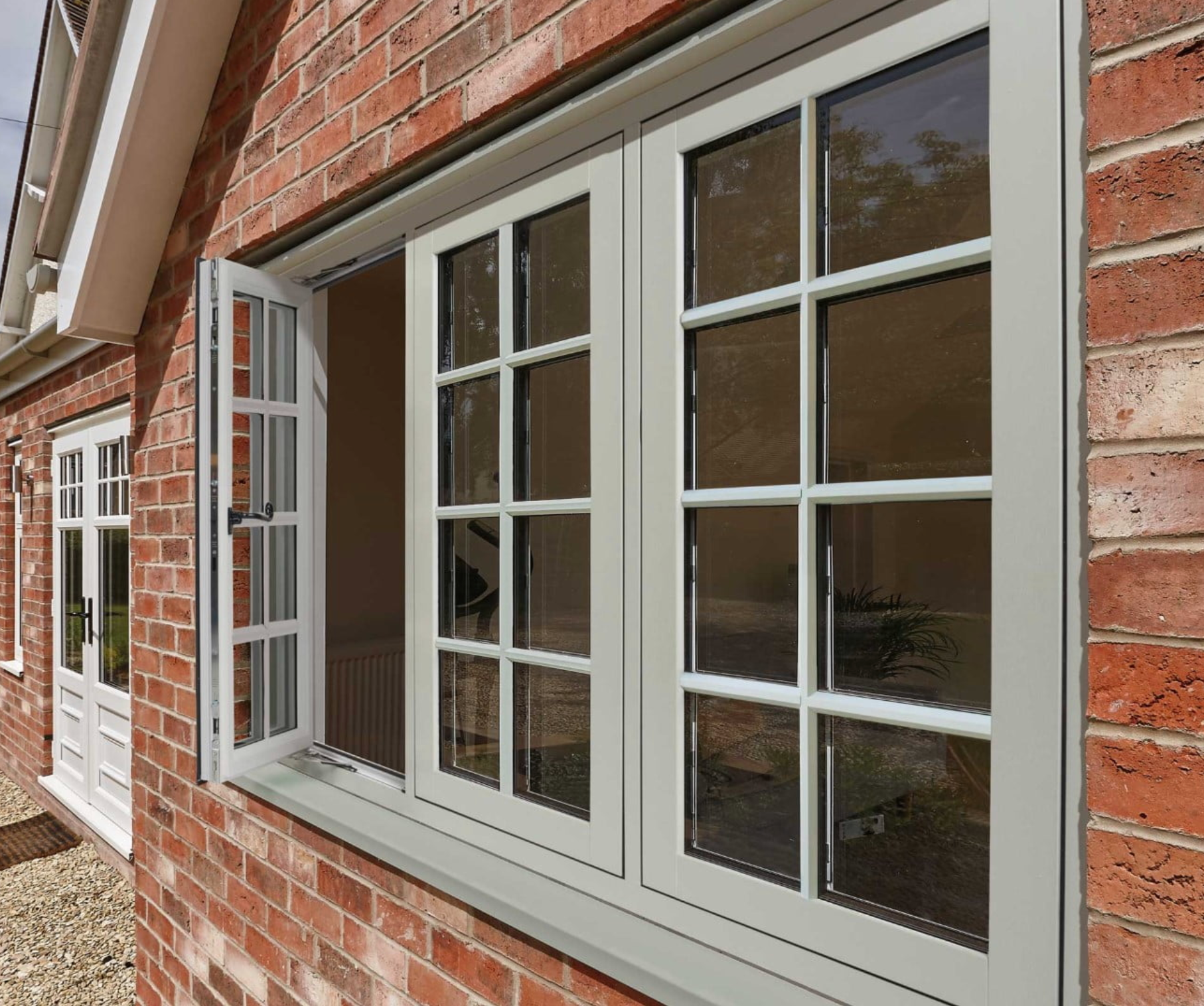 White-framed windows with a section open on a brick building, reflecting trees. Another white door to the left.