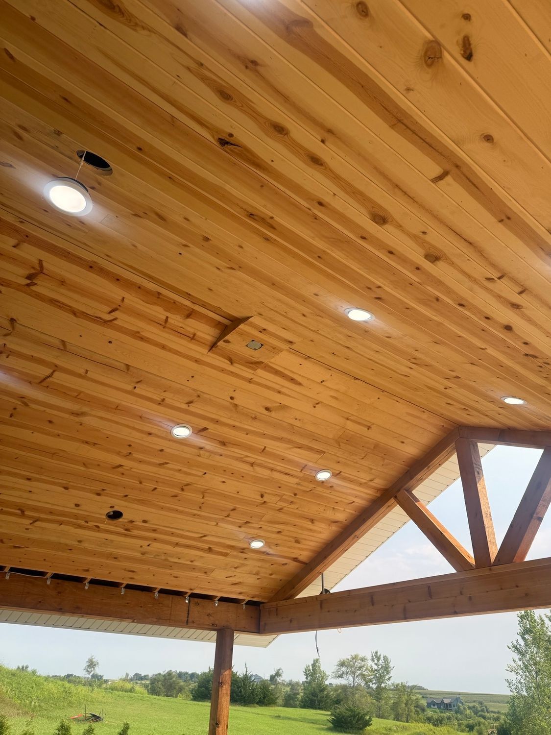 Wood-paneled pavilion ceiling with recessed lights and exposed beams, overlooking a grassy landscape.