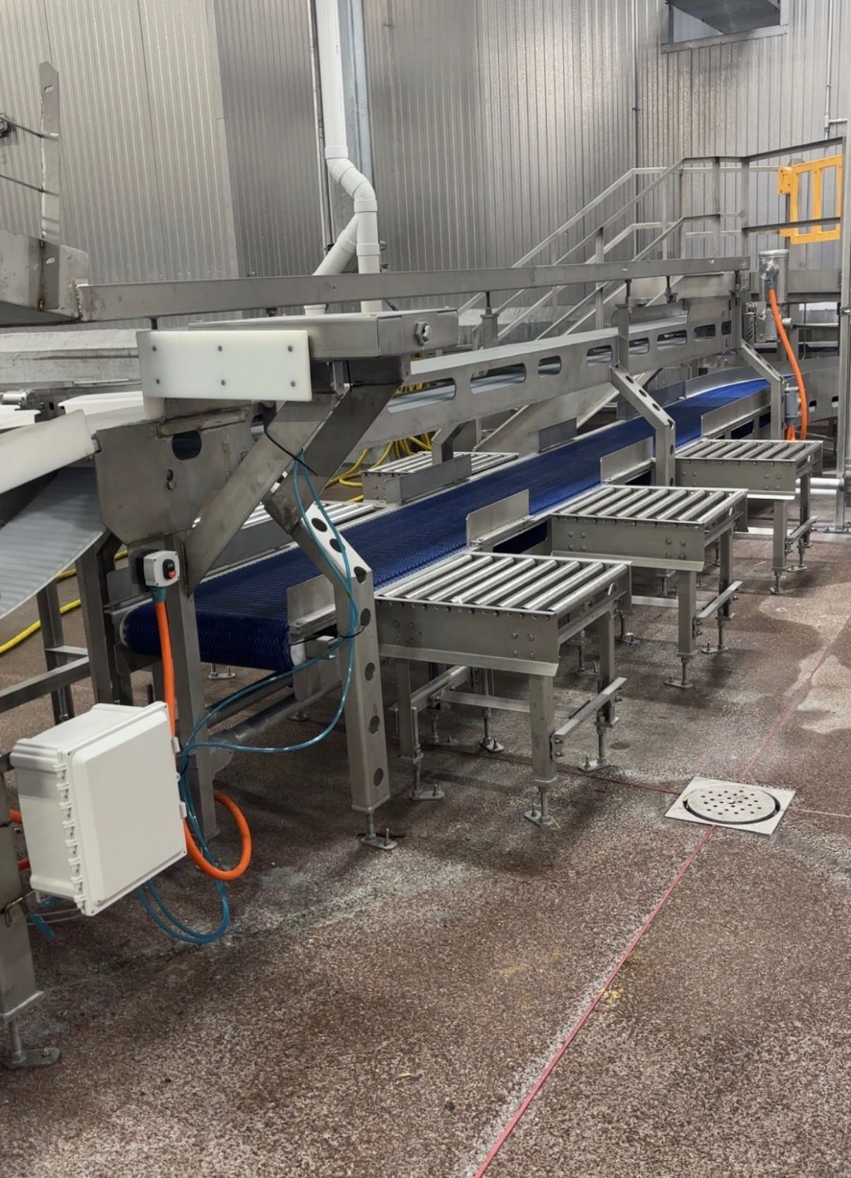 Industrial conveyor line with stainless steel platforms, blue frame, and control box in a factory setting