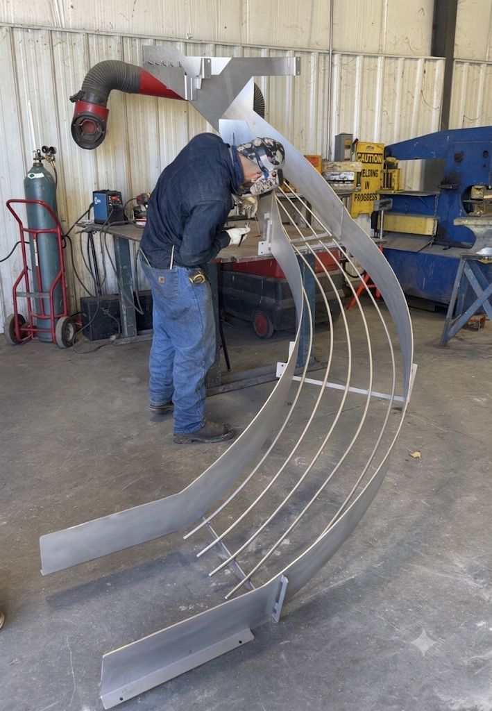 Person welding a curved metal frame in a workshop, with tools and equipment nearby.