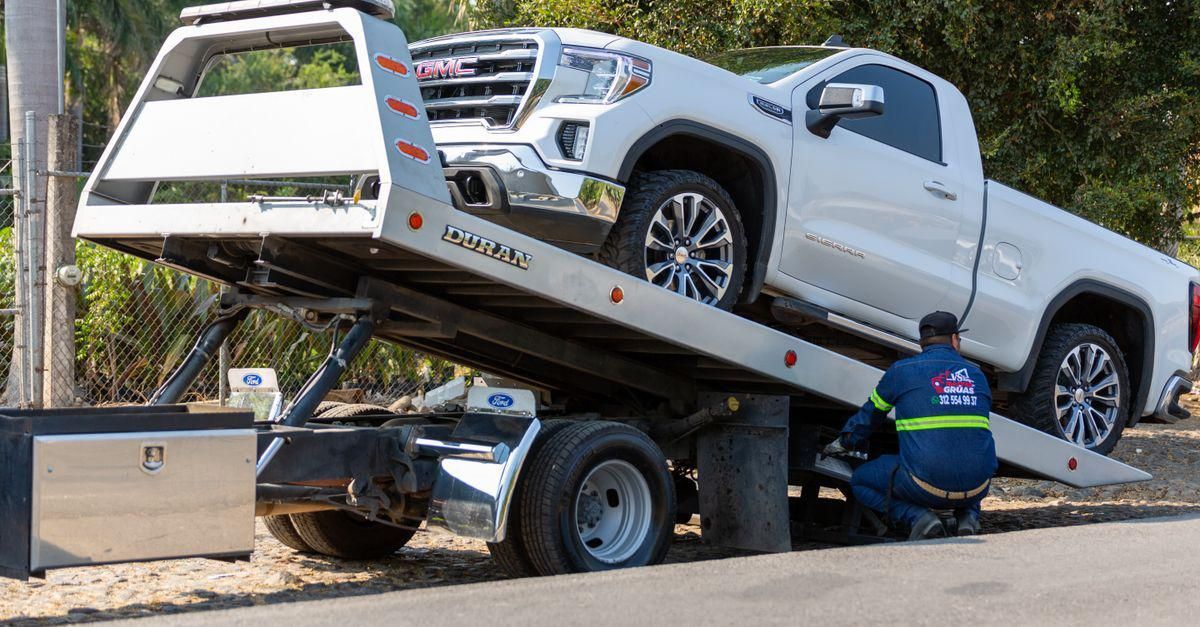 Una grúa de remolque carga una camioneta blanca en una plataforma mientras un trabajador se agacha a su lado.