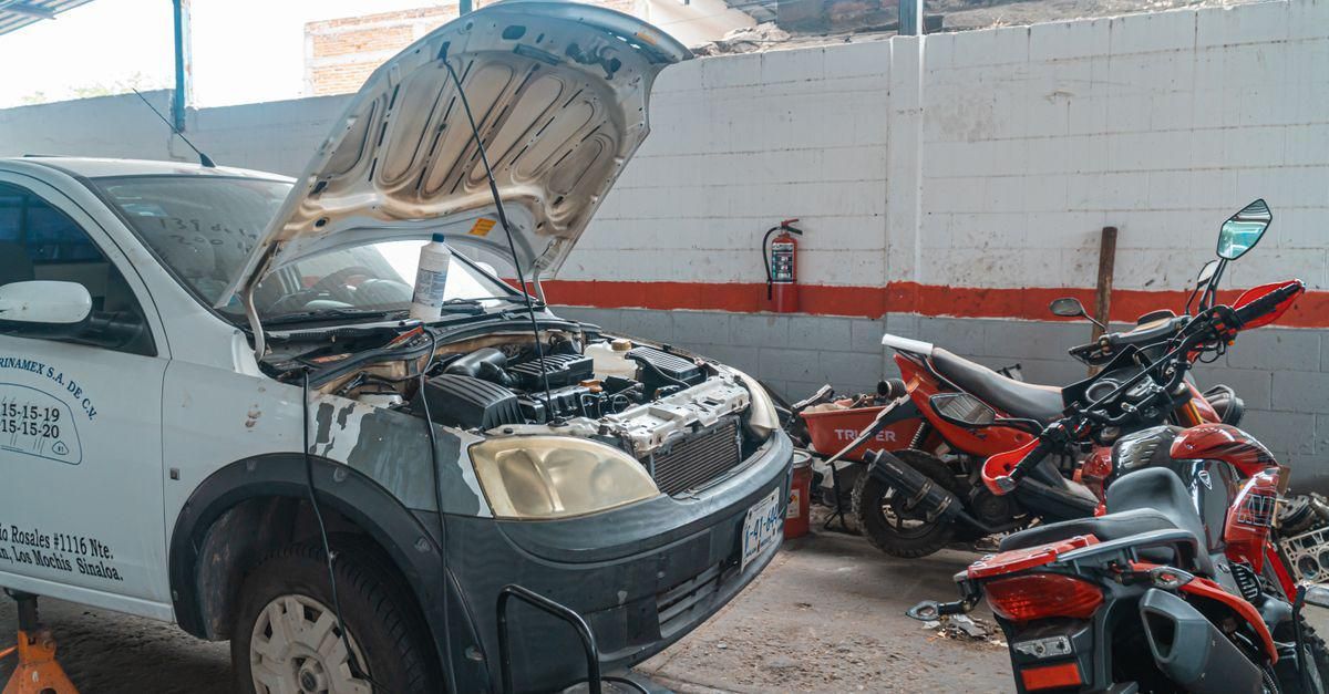 Coche gris con el capó abierto junto a varias motocicletas rojas en un garaje o taller de reparación