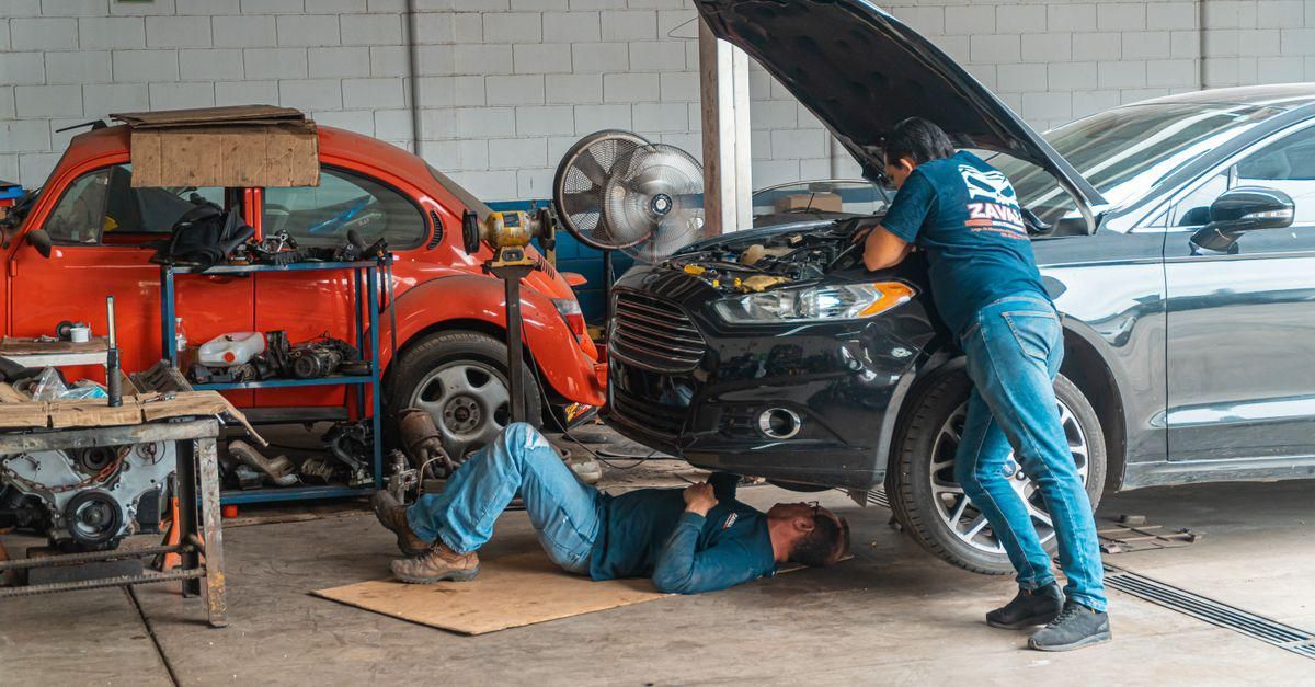 Mecánicos reparando coches en un taller, con un vehículo rojo y otro gris en reparación
