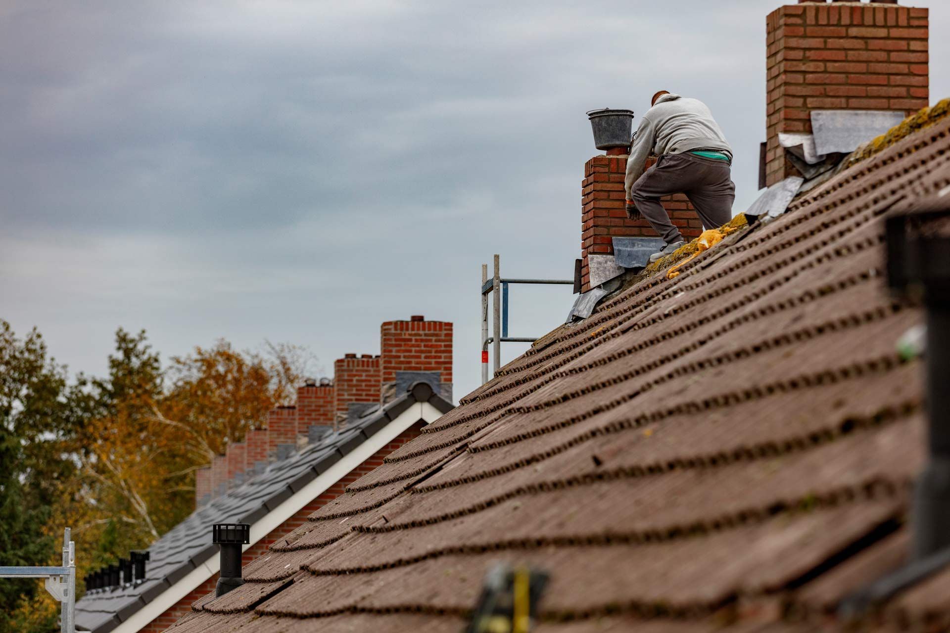 Roofer working on a sloped shingle roof beside brick chimneys under a cloudy sky