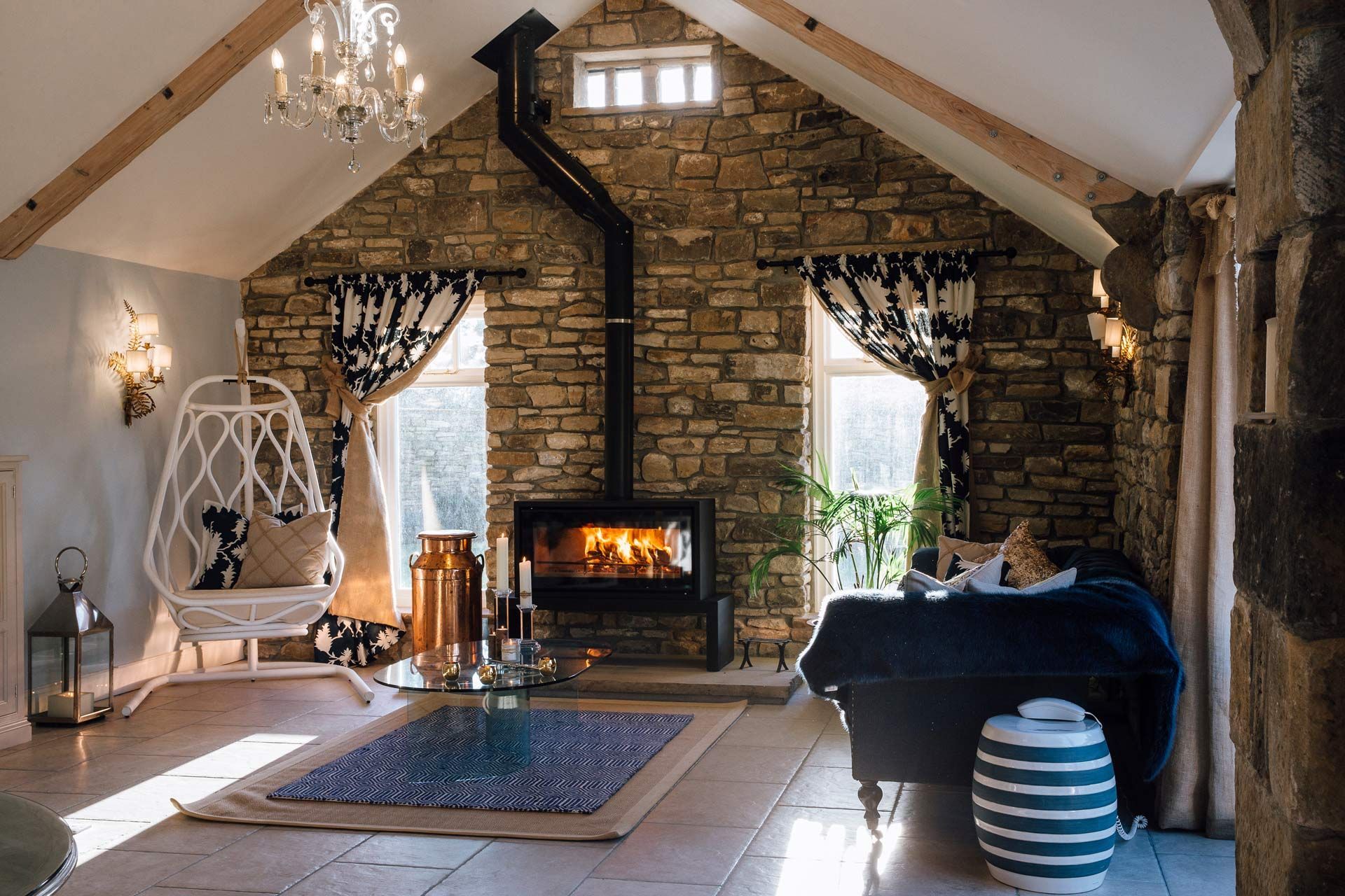 Cozy rustic living room with stone fireplace, chandelier, blue sofa, and hanging chair by bright windows