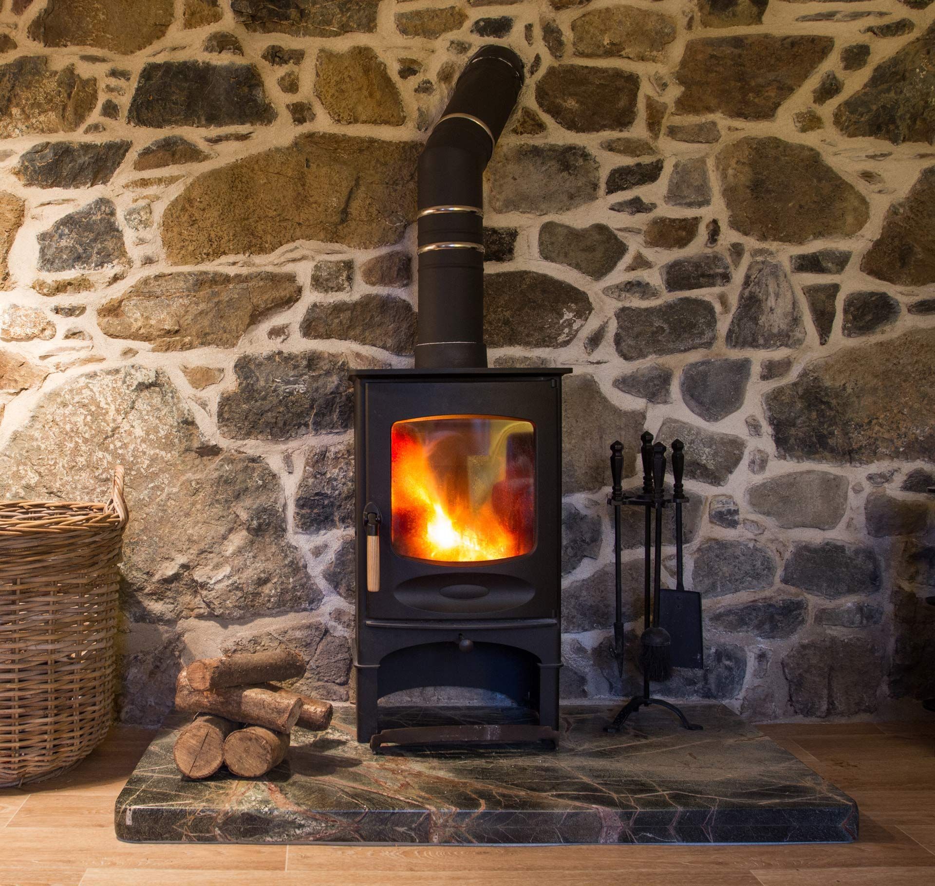 Wood-burning stove with bright fire against a stone wall, beside logs and fireplace tools