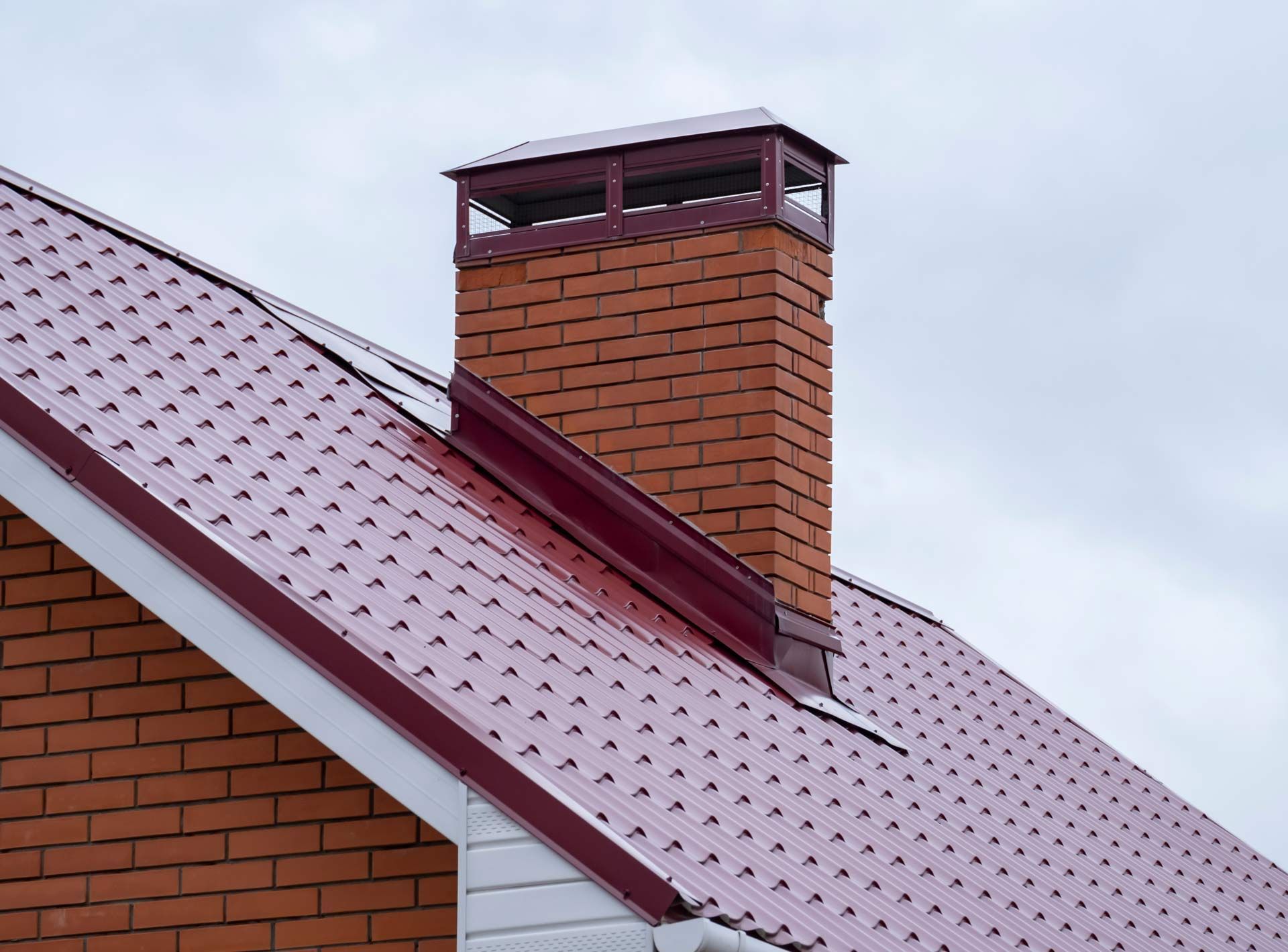 Brick chimney with a metal cap on a red-roofed house under a cloudy sky