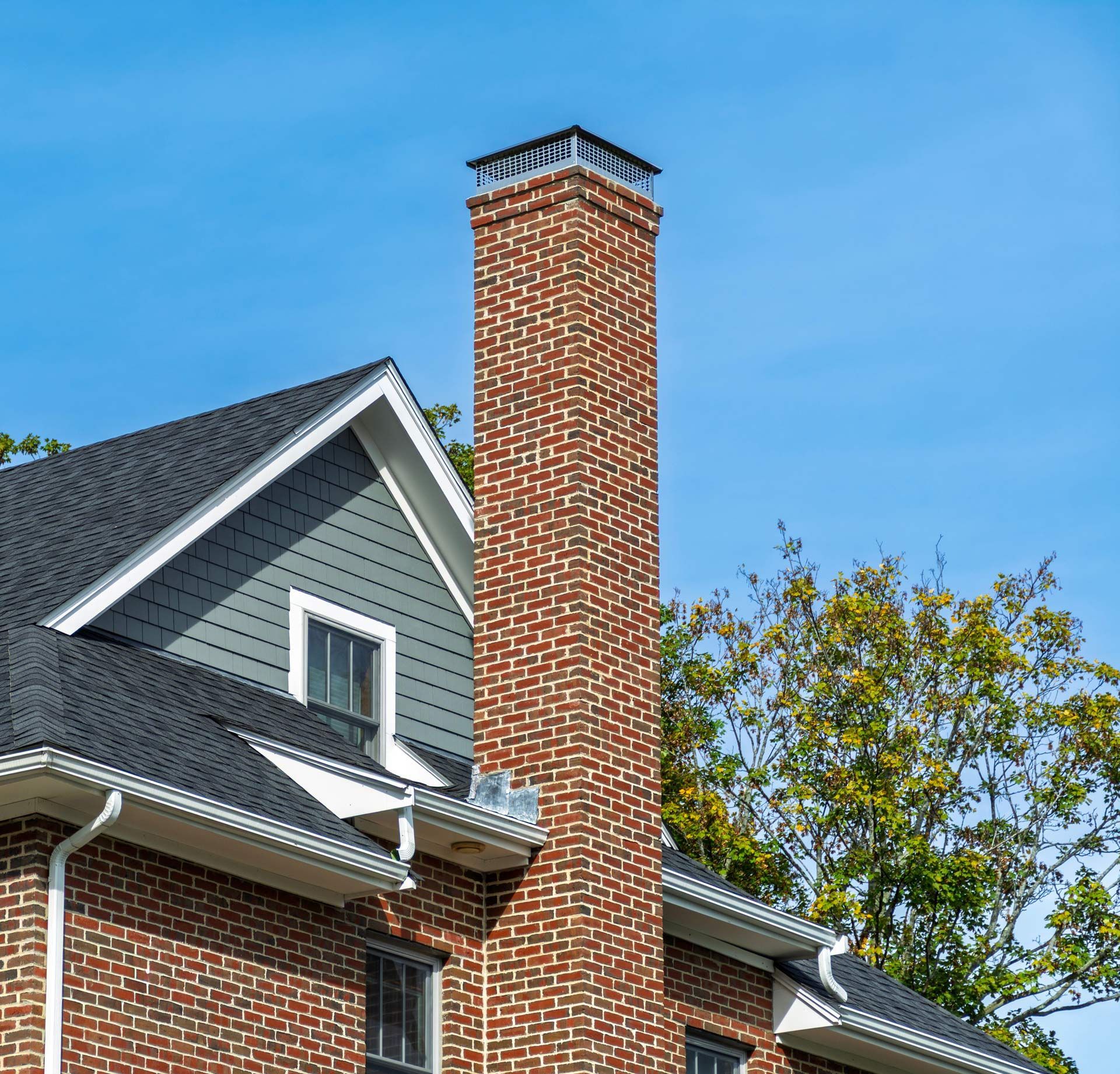 Brick chimney on a gray house roof against a blue sky and trees