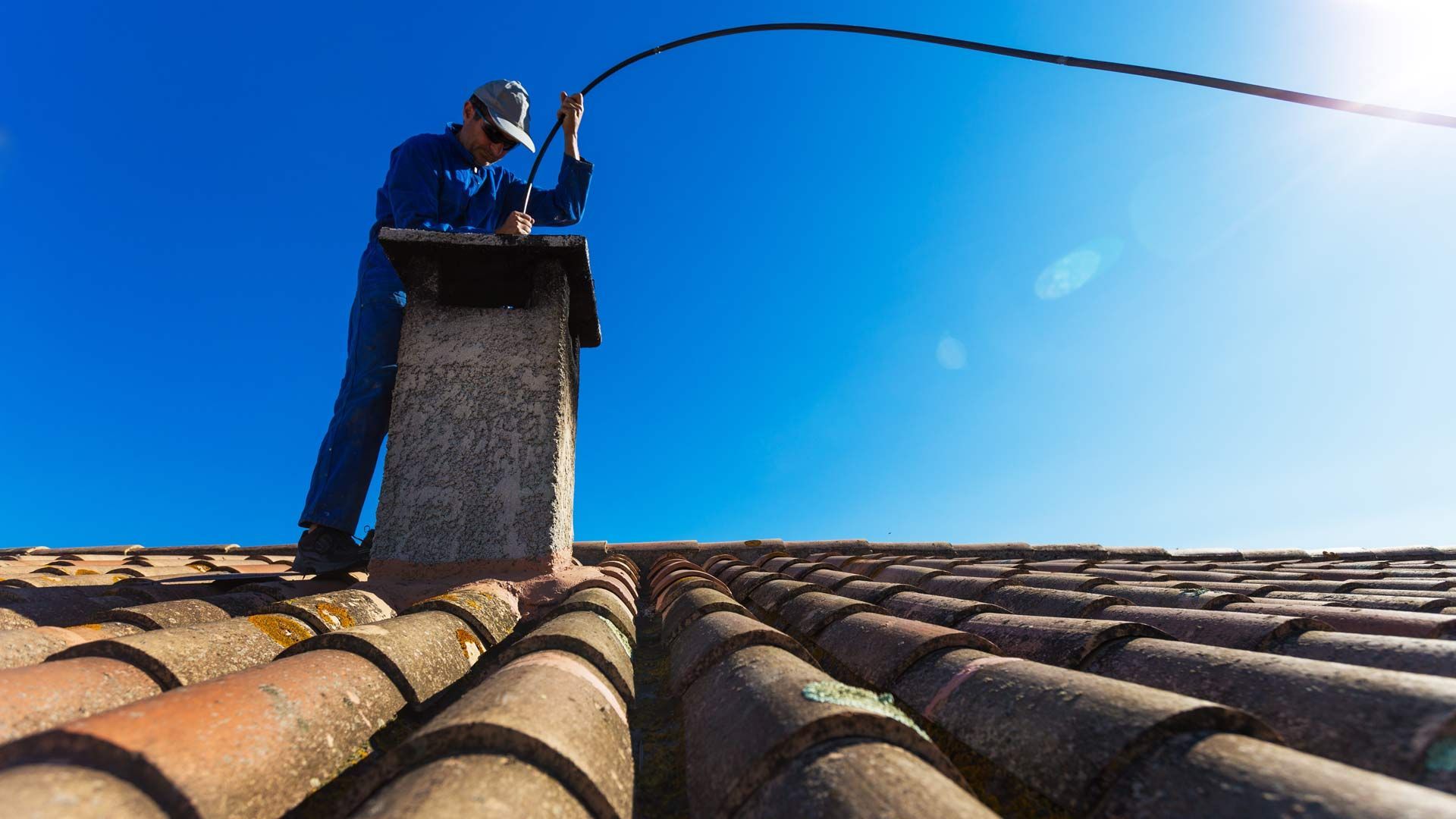Roofing worker standing on a chimney, sweeping soot across a terracotta-tiled roof under a clear blue sky