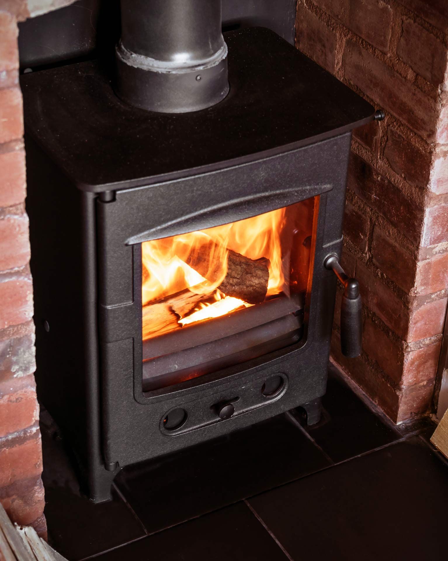 Black wood-burning stove with bright fire inside, set in a brick corner on a tiled floor.