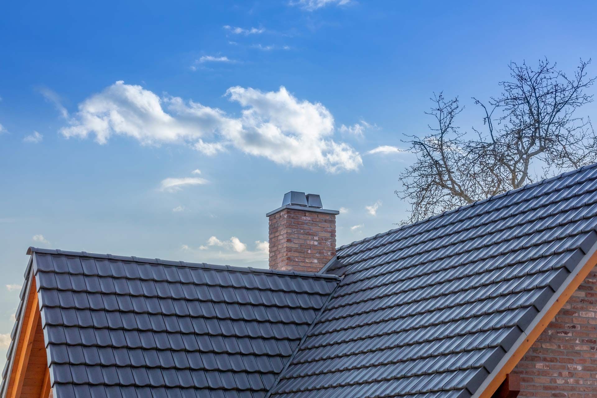 Dark gray tiled roof with a brick chimney under a blue sky with clouds