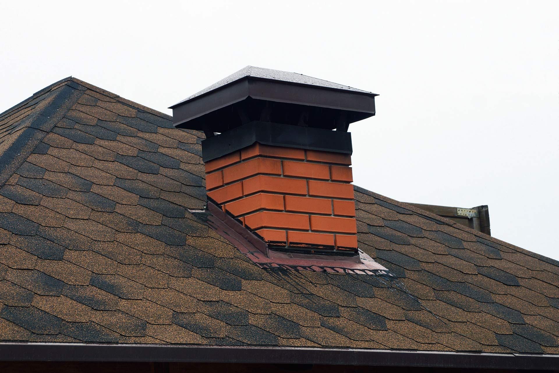 Rooftop chimney with red bricks and a black cap on a shingled roof under a cloudy sky