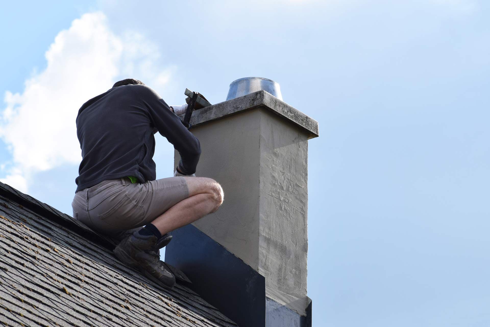 Person crouching on a sloped roof beside a chimney under a blue sky