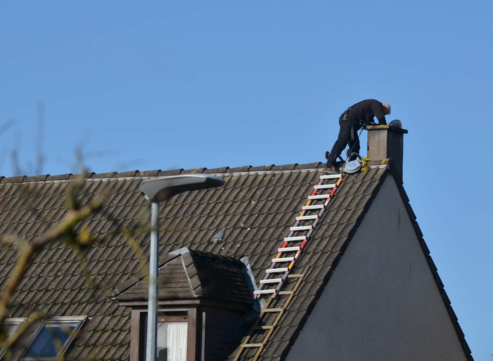 Worker on a roof near a ladder, repairing shingles under a clear blue sky.