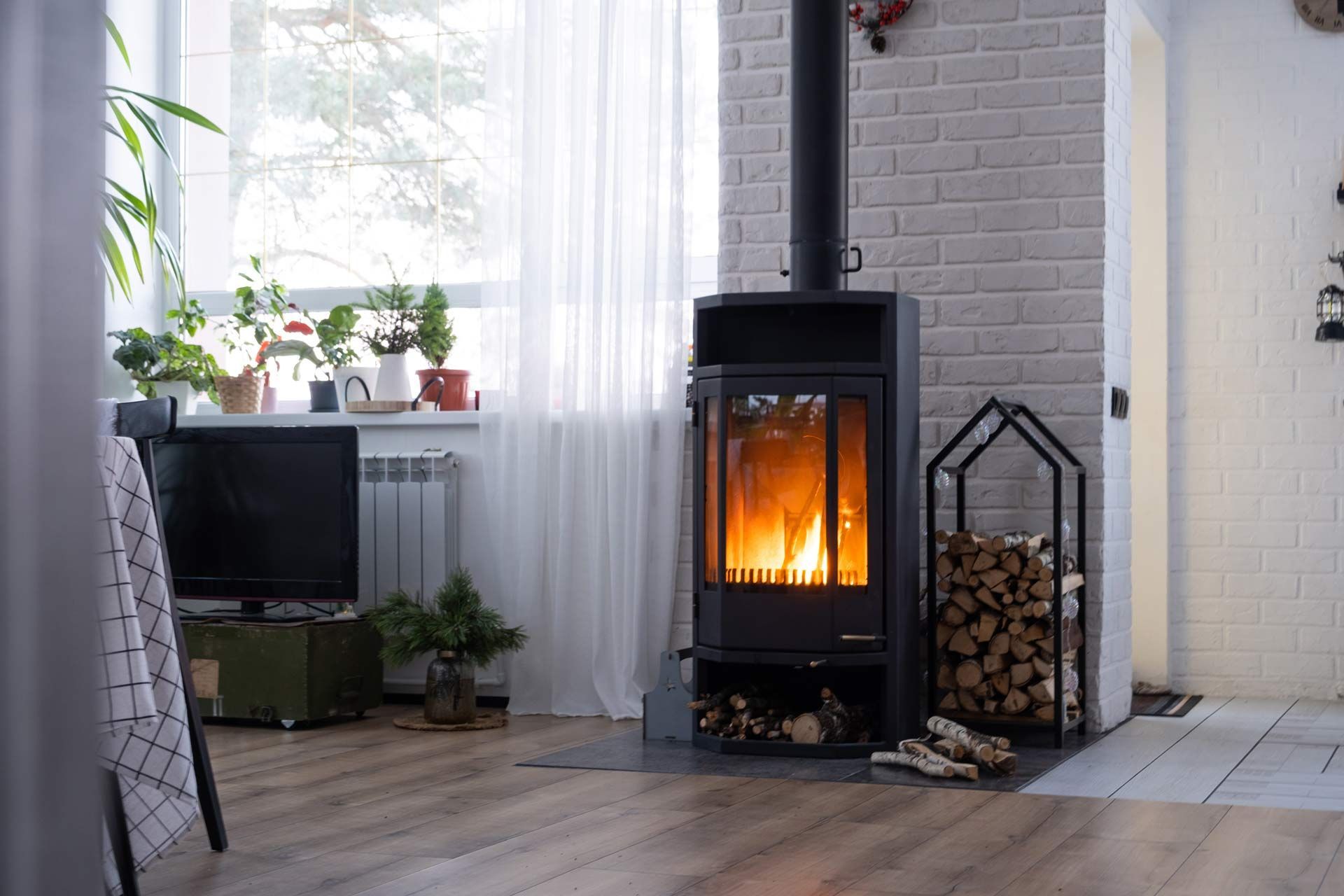 Cozy living room with a lit black wood stove beside a window, potted plants, and stacked firewood in a basket.