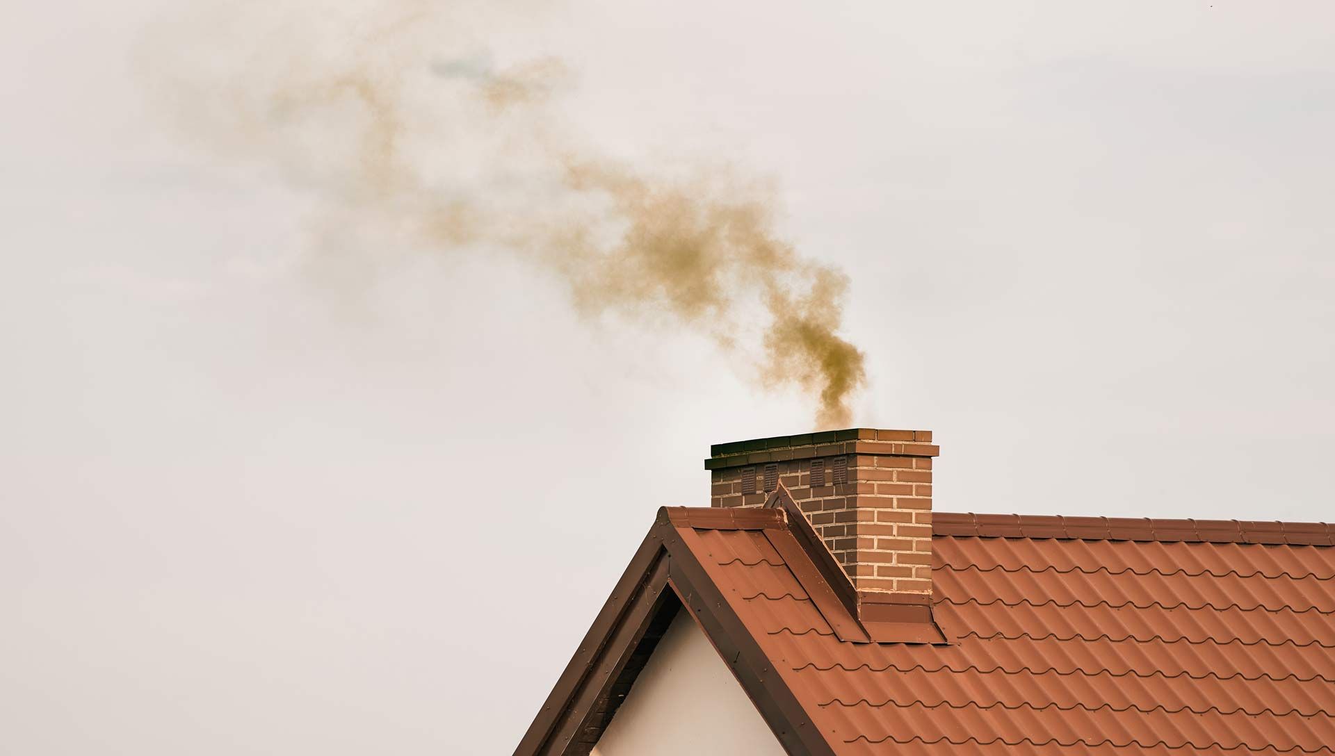 Smoky chimney on a red-roofed house against a pale sky
