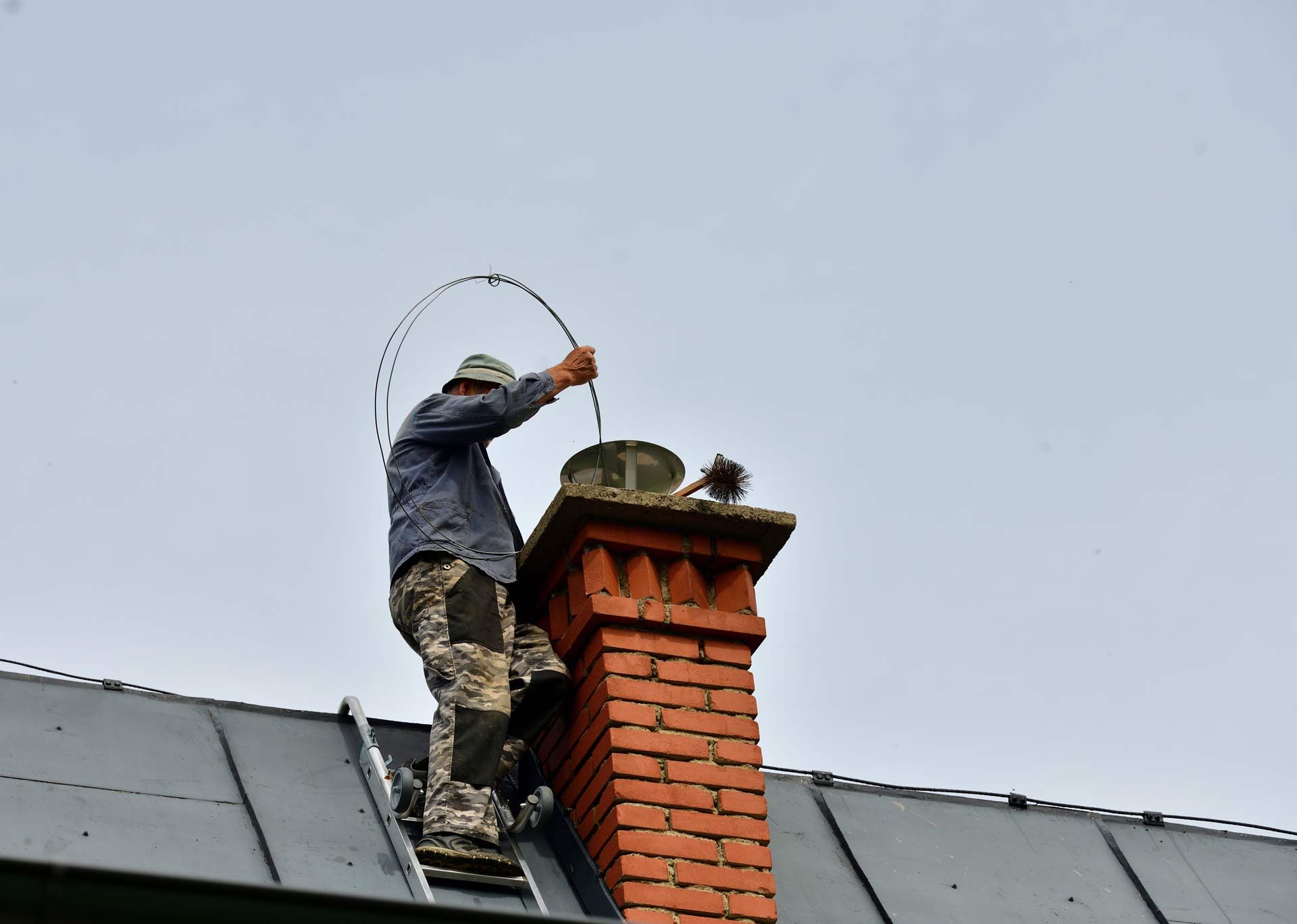 Worker on a roof cleaning a brick chimney with a brush under a cloudy sky