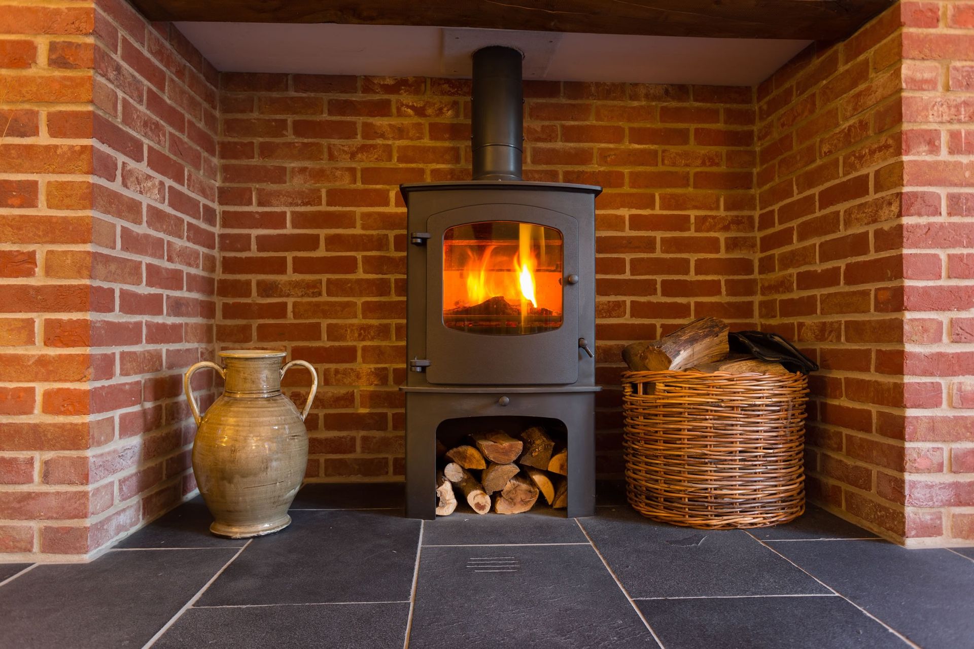 Wood-burning stove in a brick fireplace with fire, logs, ceramic jar, and wicker basket